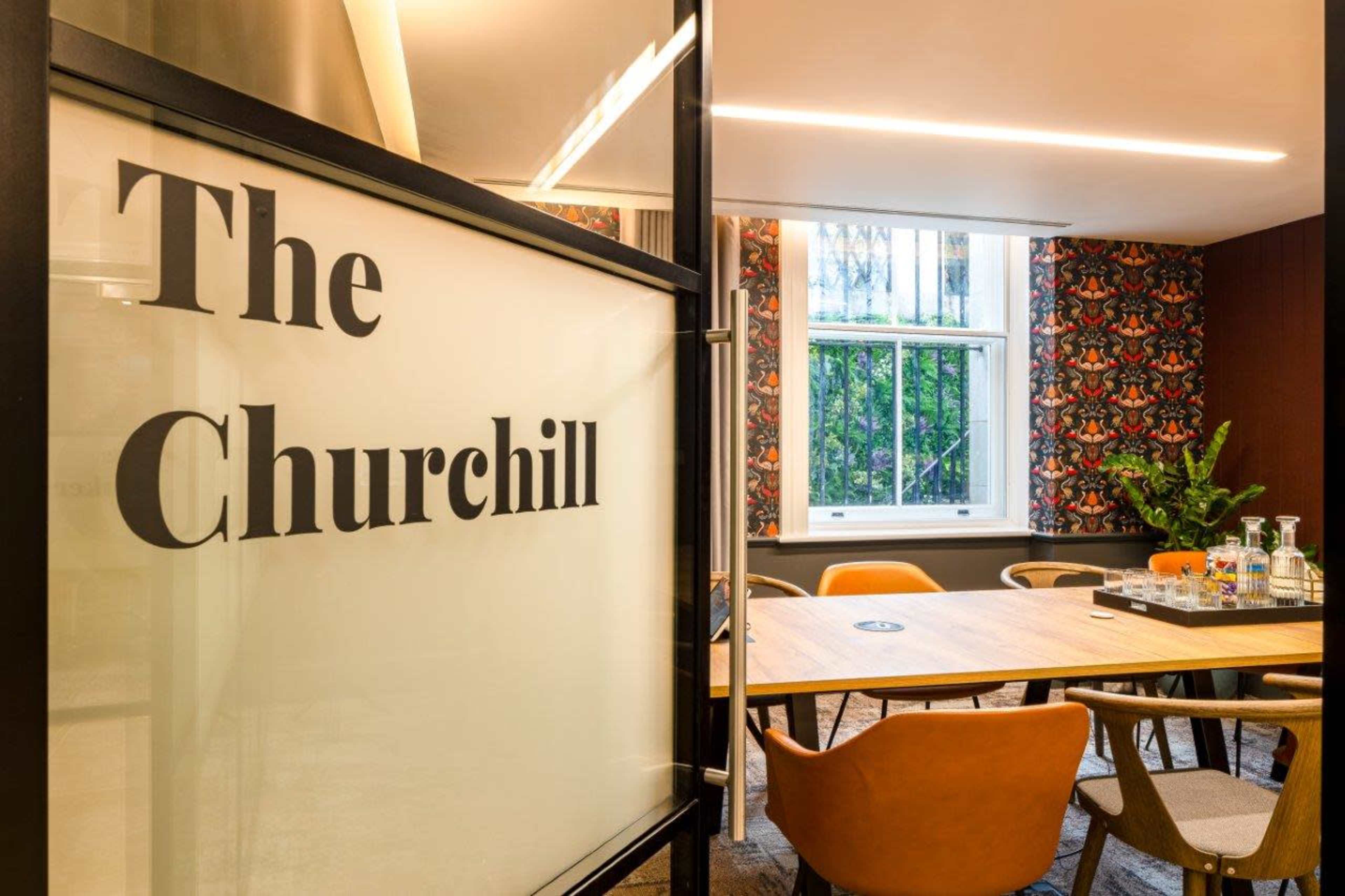 A meeting room named "The Churchill," featuring a large wooden table surrounded by chairs and a window with decorative wallpaper.