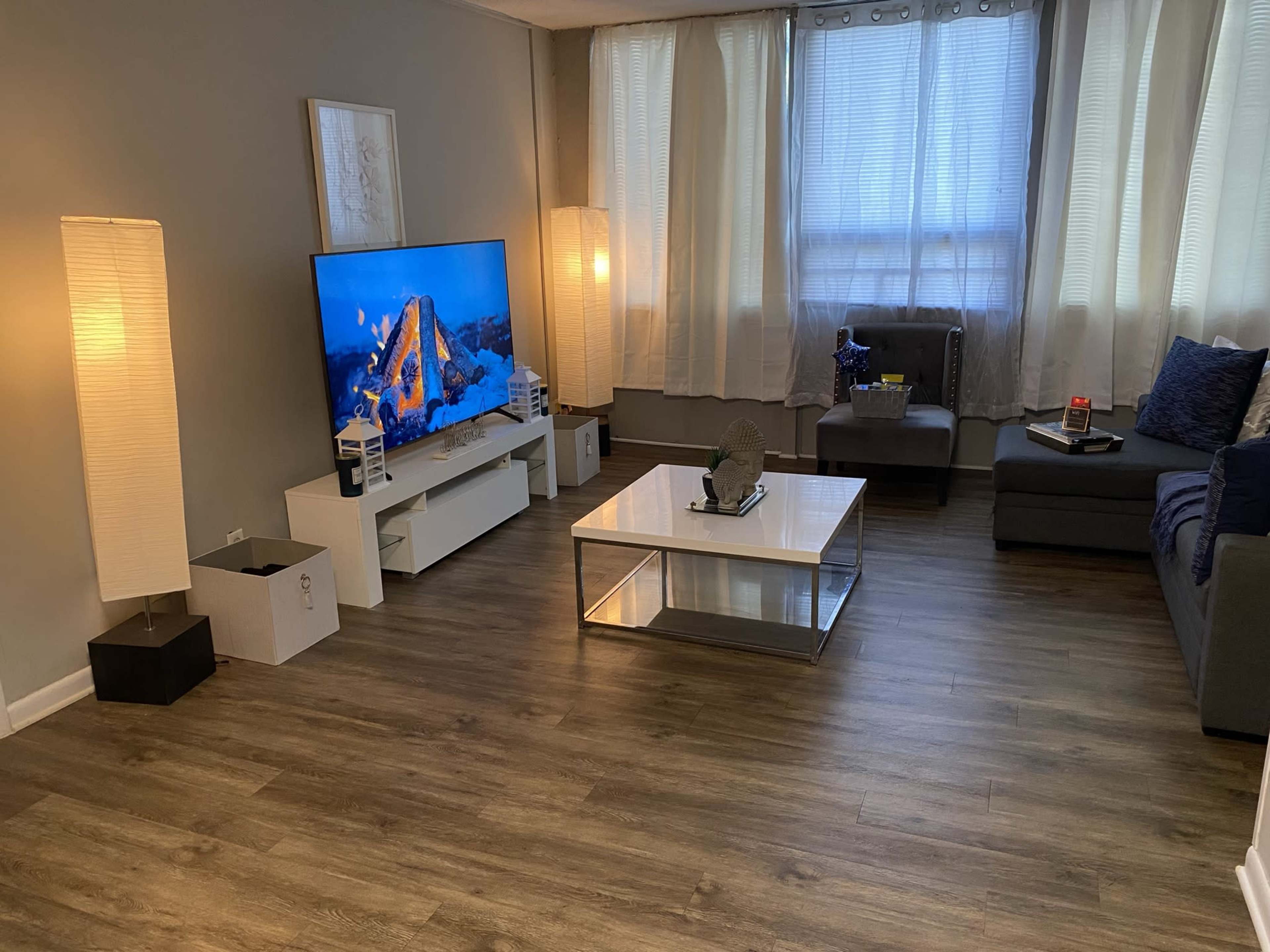 The room features a modern living area with a large television on a white cabinet, a glass coffee table, and a gray sofa, illuminated by two floor lamps and natural light from large windows.