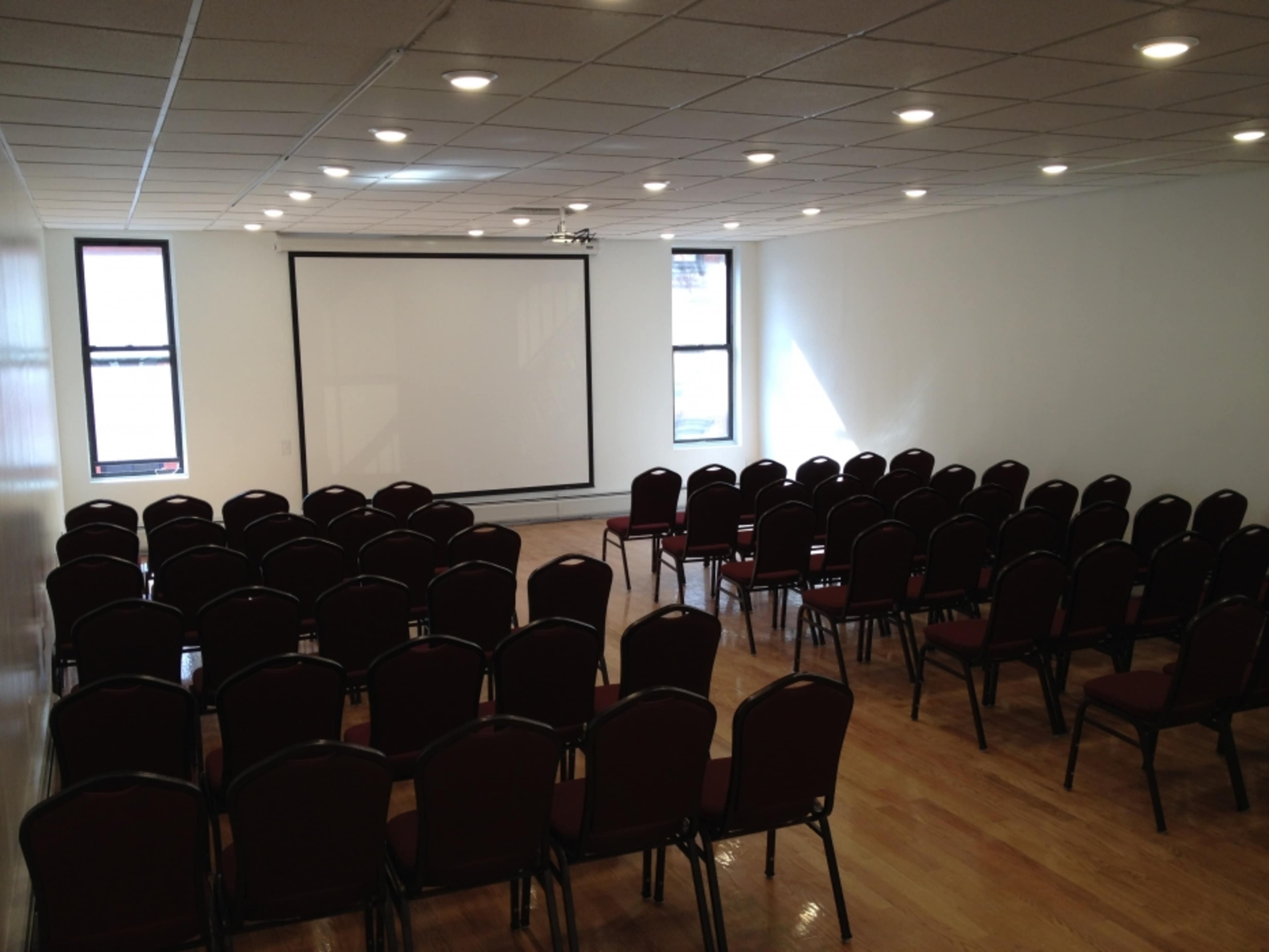 The image shows an empty conference room with rows of red chairs facing a projection screen.