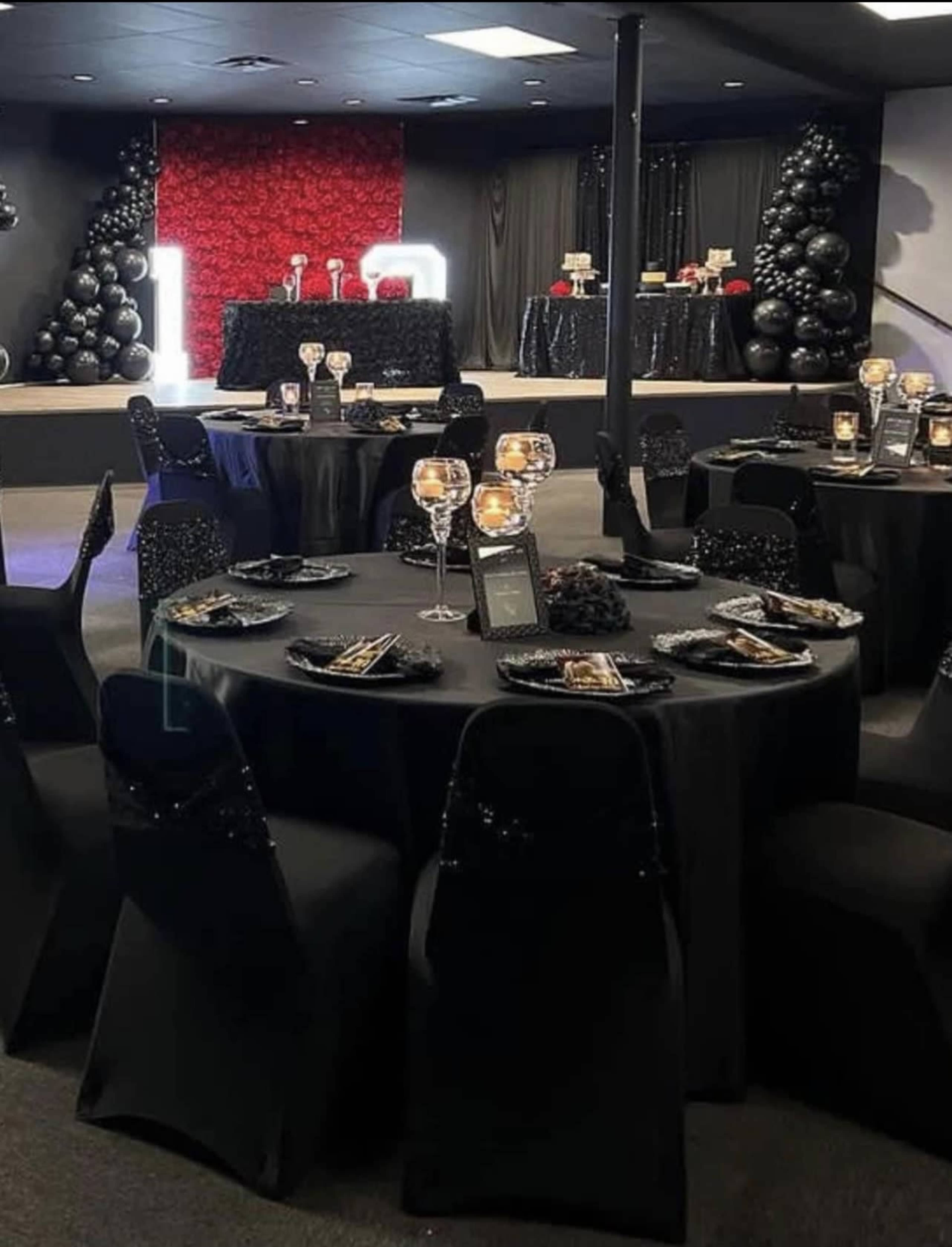 The image shows a formal dining setup with black tables and chairs, decorated with elegant tableware, centered around a backdrop of red roses and black decor elements.