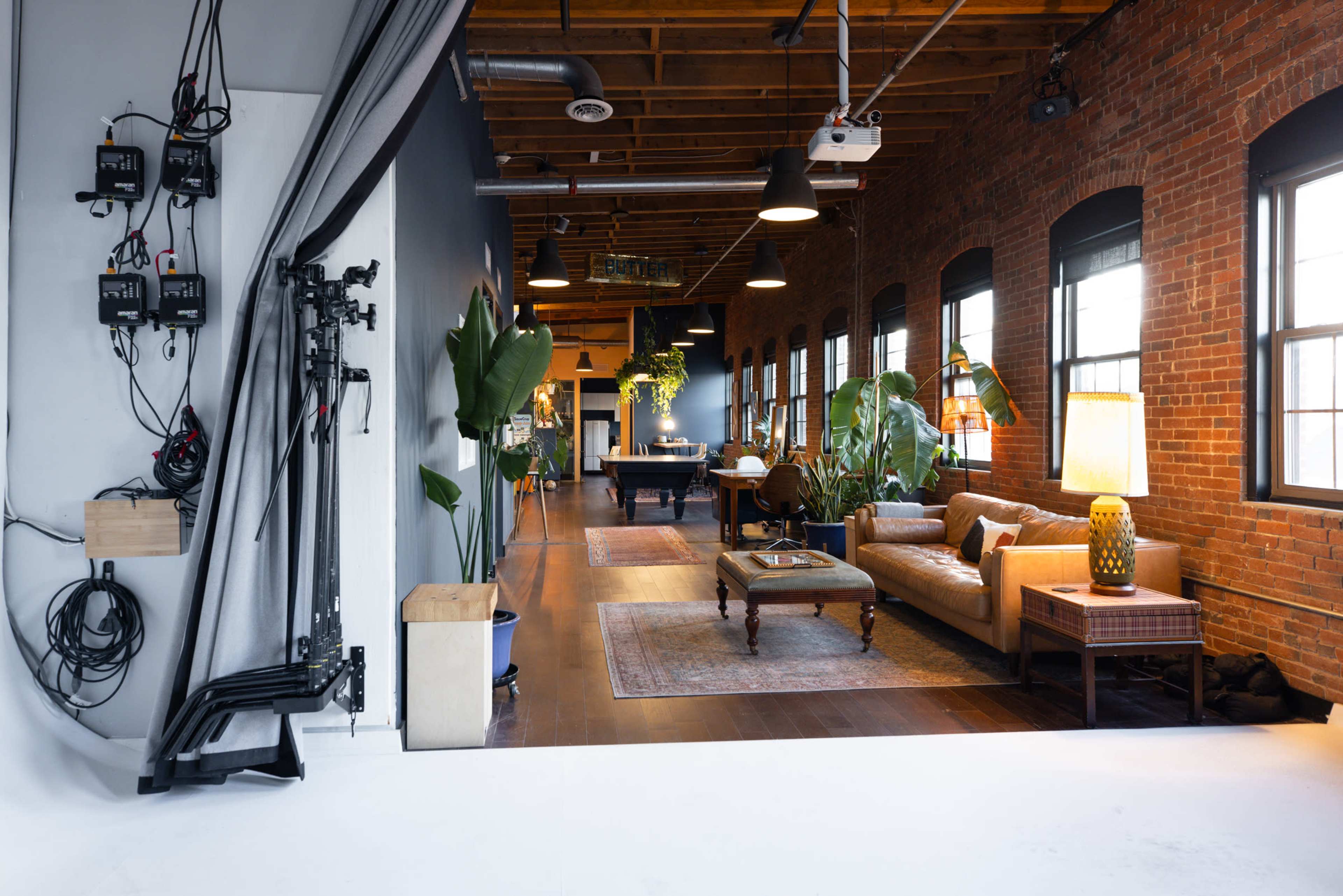 The image depicts an industrial-style interior space with brick walls, wooden beams, and modern furniture, including a couch and a pool table, alongside plants and decorative lighting.