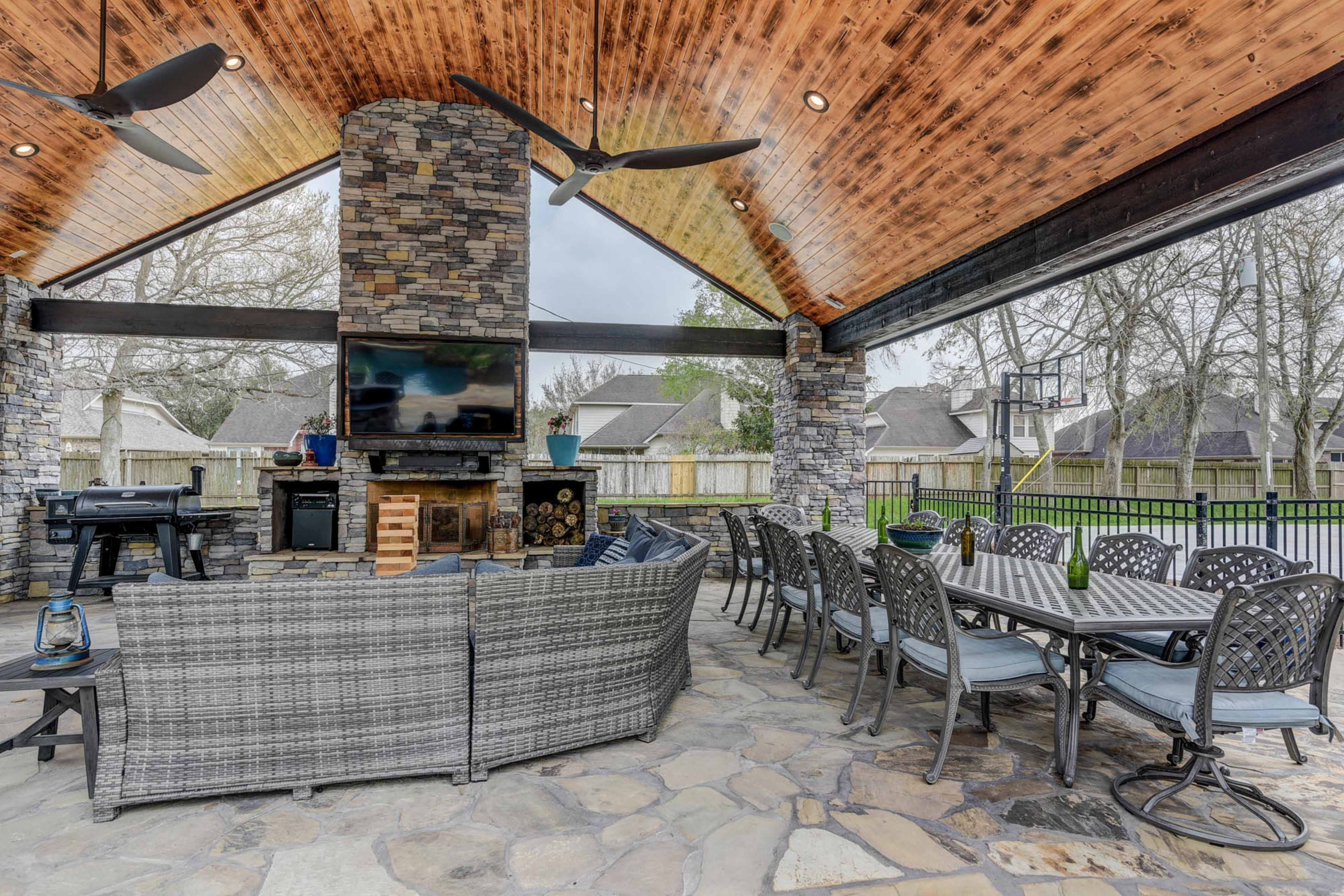 A spacious outdoor patio area featuring stone walls, a large stone fireplace, a dining table, and a barbecue grill under a high ceiling.