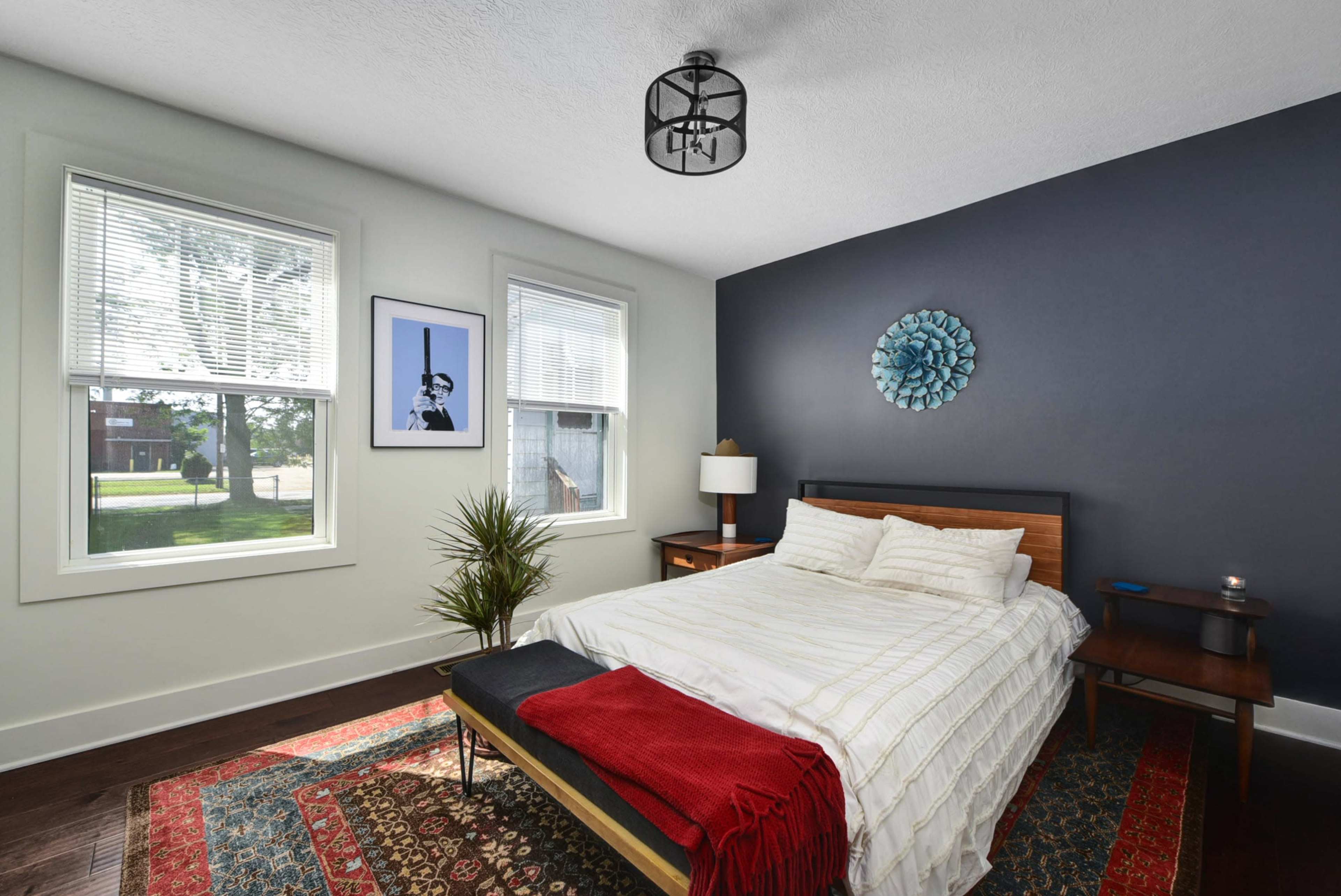 A neatly arranged bedroom with a bed, two windows, a plant, and a decorative wall piece on a dark-colored wall.