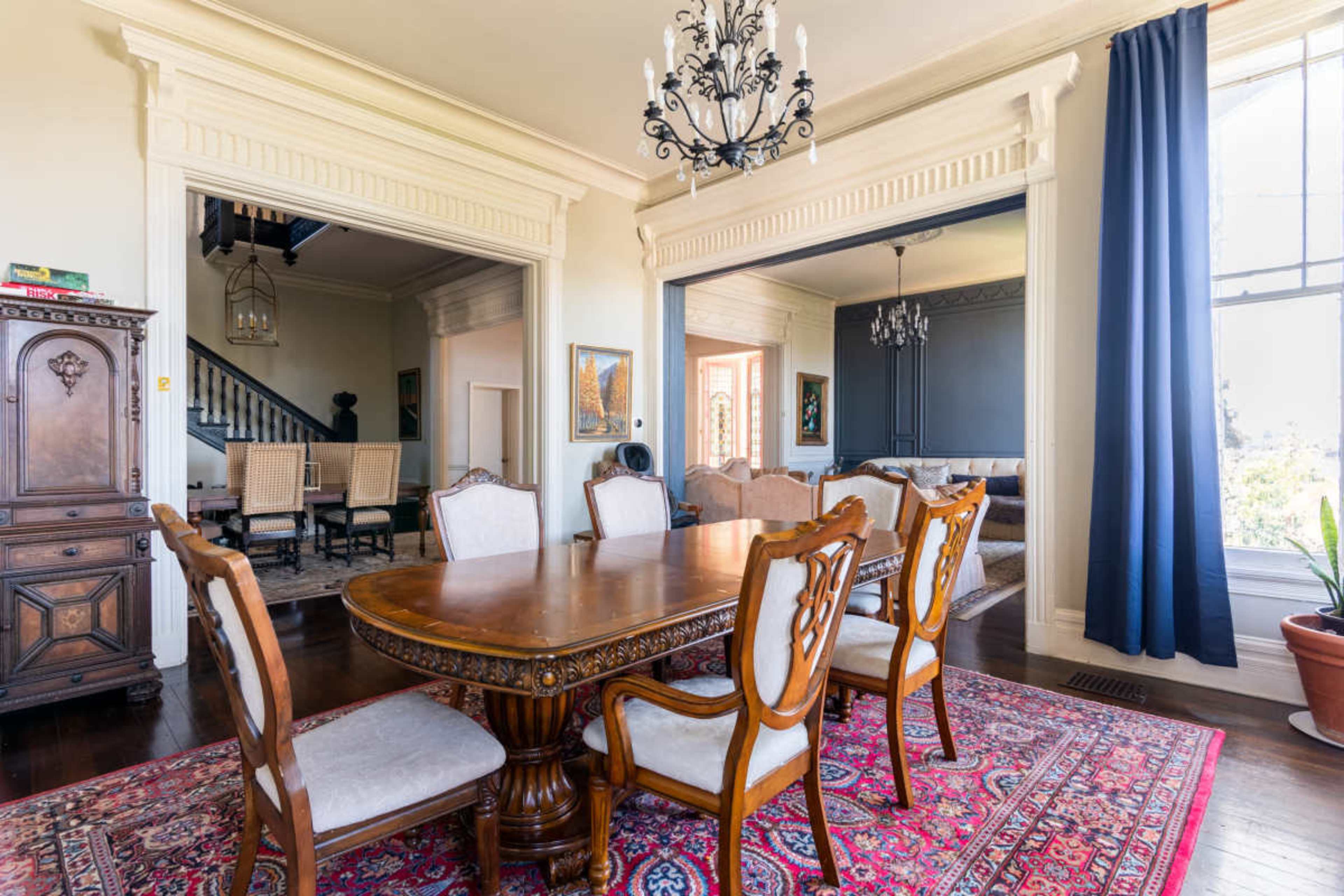 The image depicts a dining room with a wooden table surrounded by upholstered chairs, set against a backdrop of elegant architectural features and large windows.