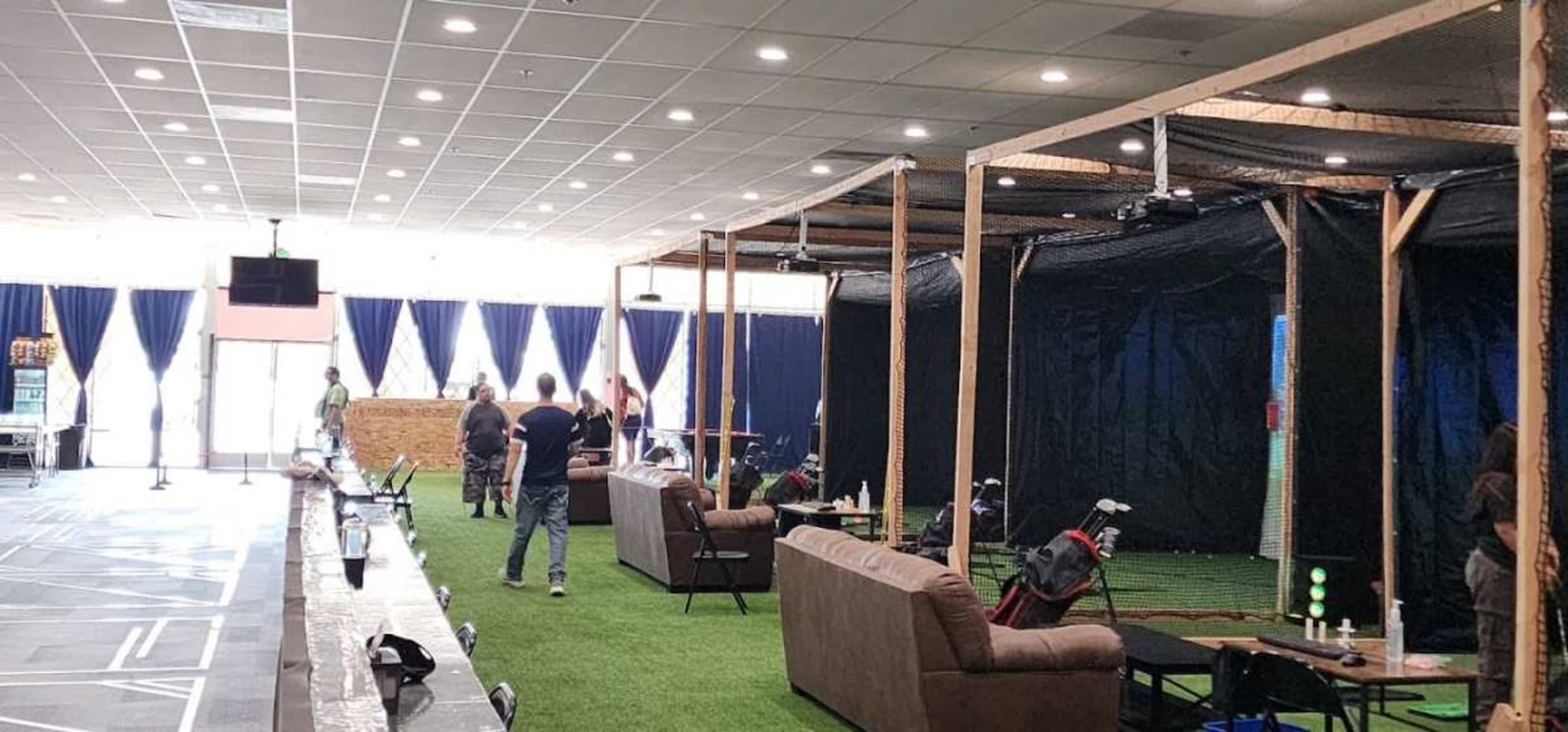 The image shows an indoor golf facility with a grassy area, seating, and several people engaged in activities.