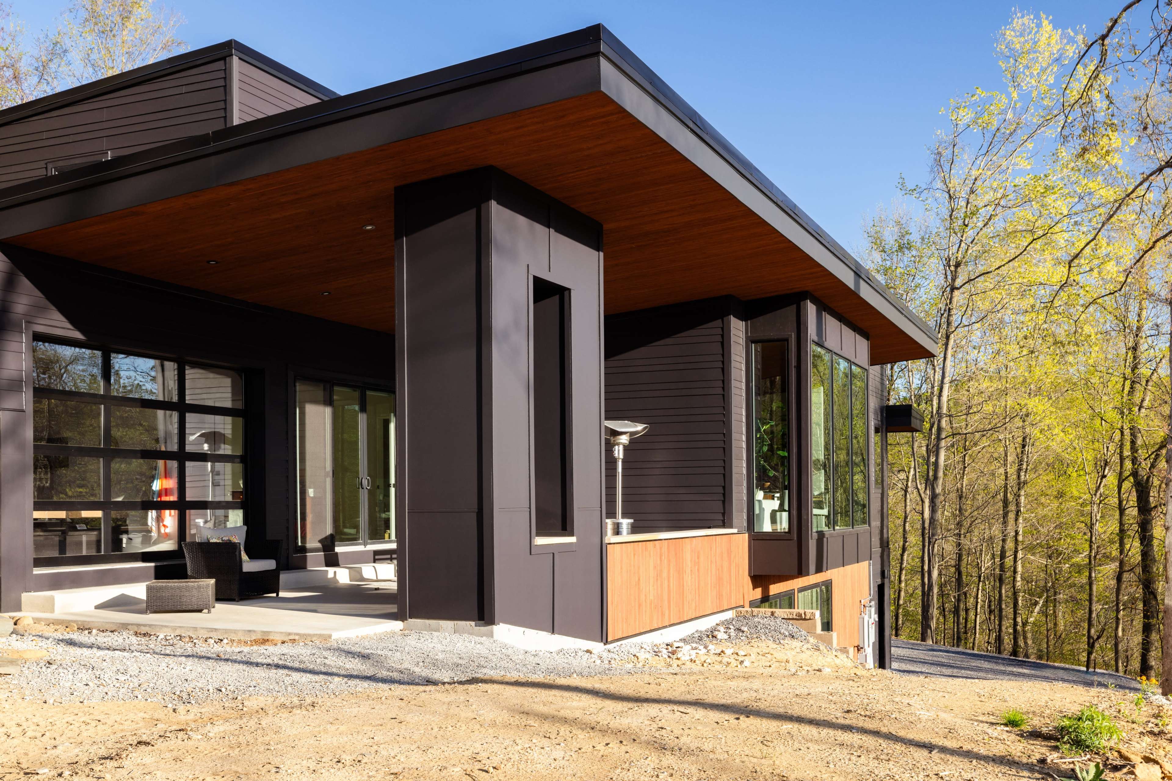 The image shows a modern house with dark exterior walls and large windows, surrounded by trees and a gravel pathway.