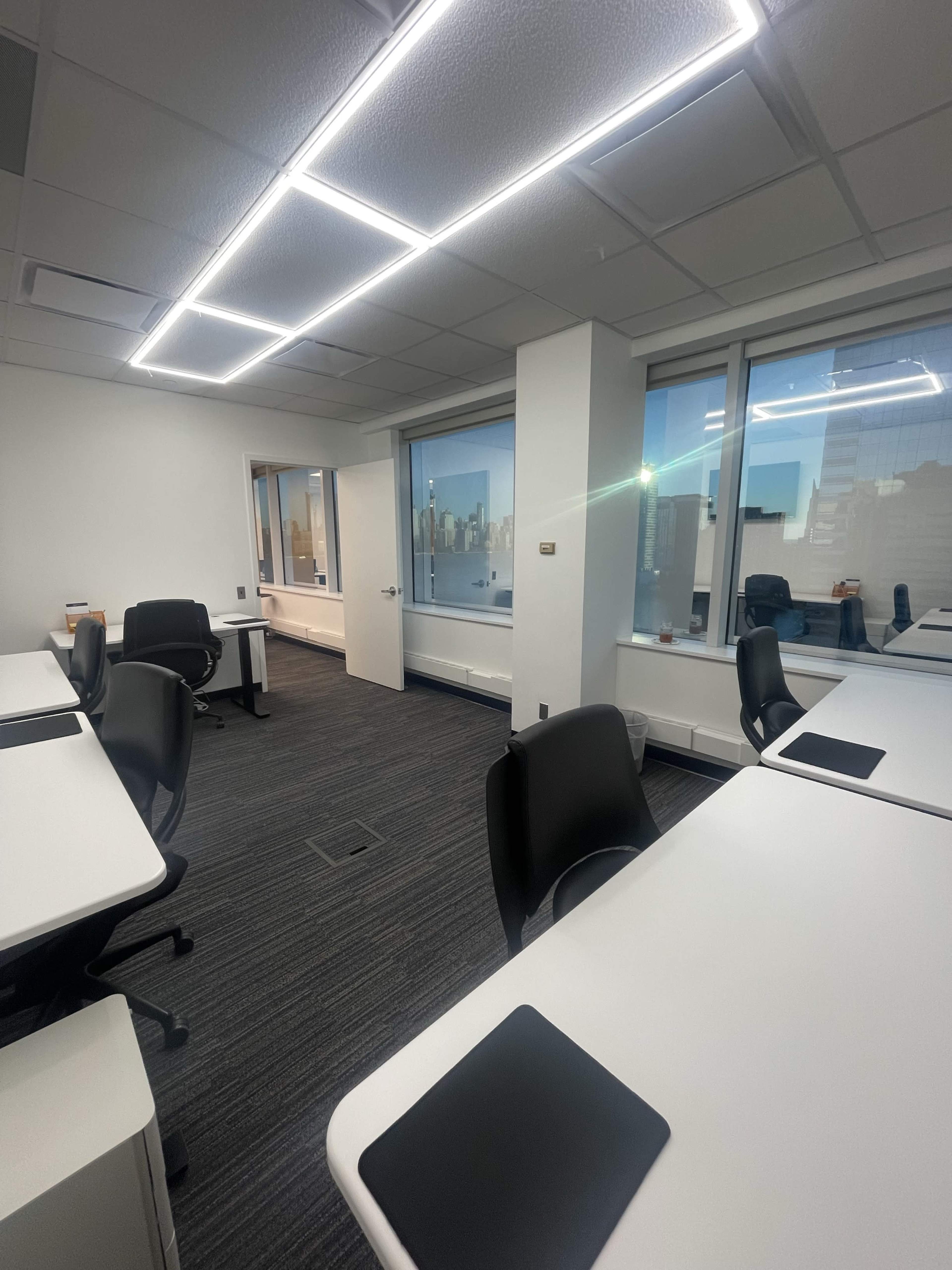 A modern office space with white desks and black chairs, featuring large windows overlooking a city skyline.
