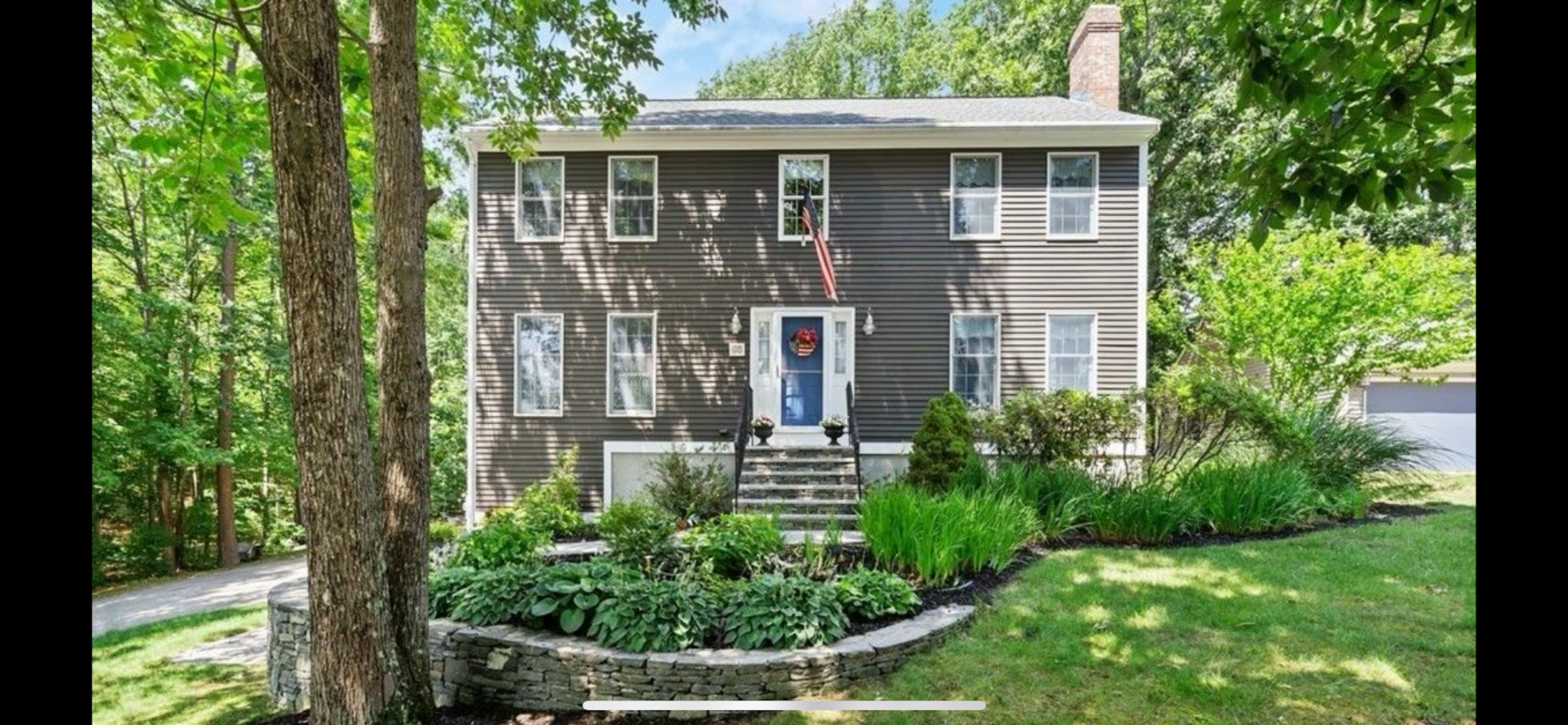 The image shows a two-story brown house with white shutters and a blue front door, surrounded by a landscaped yard with trees and shrubs.