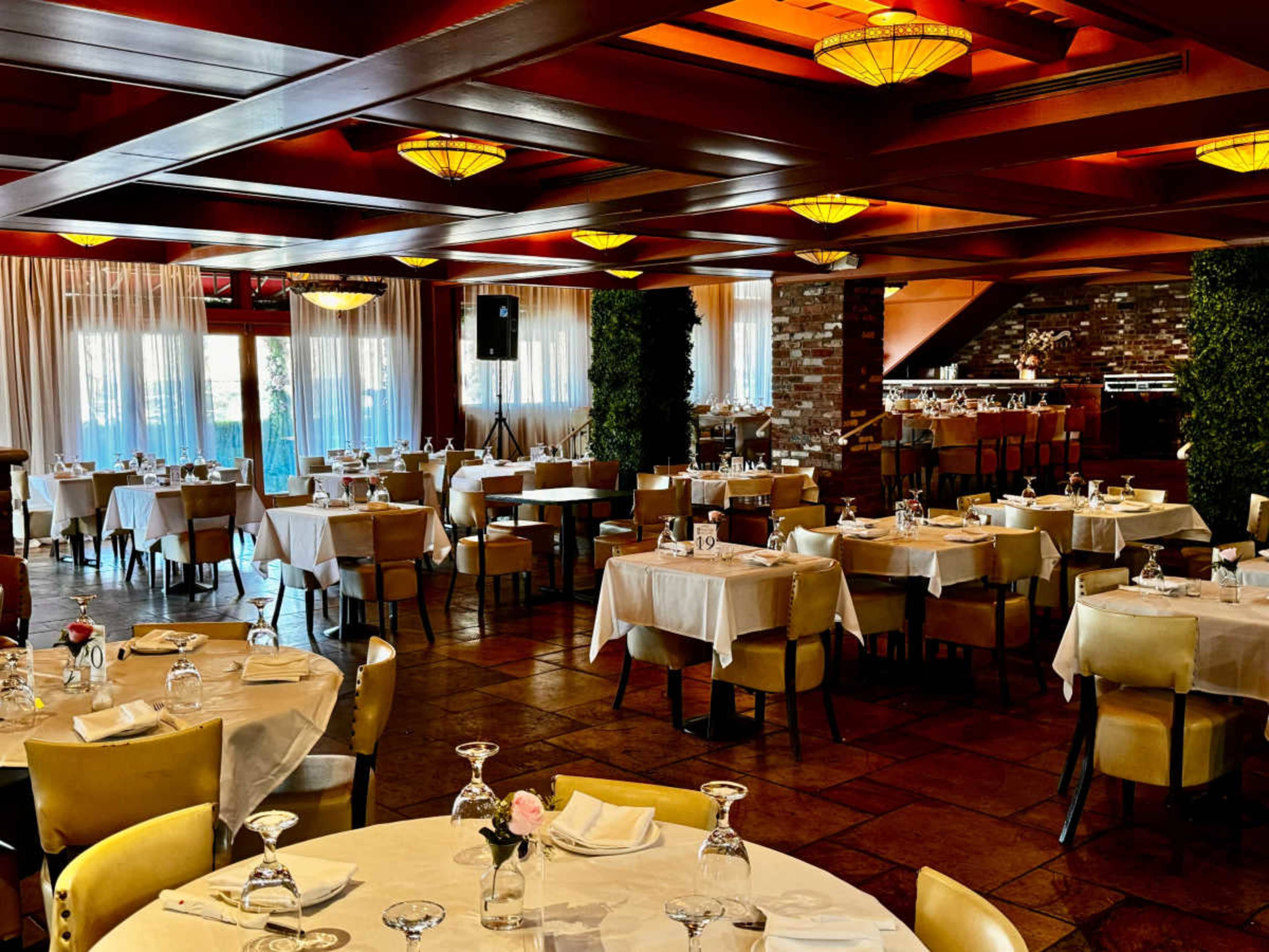 The image shows a spacious restaurant interior with multiple dining tables set with glassware and white tablecloths, illuminated by pendant lights.