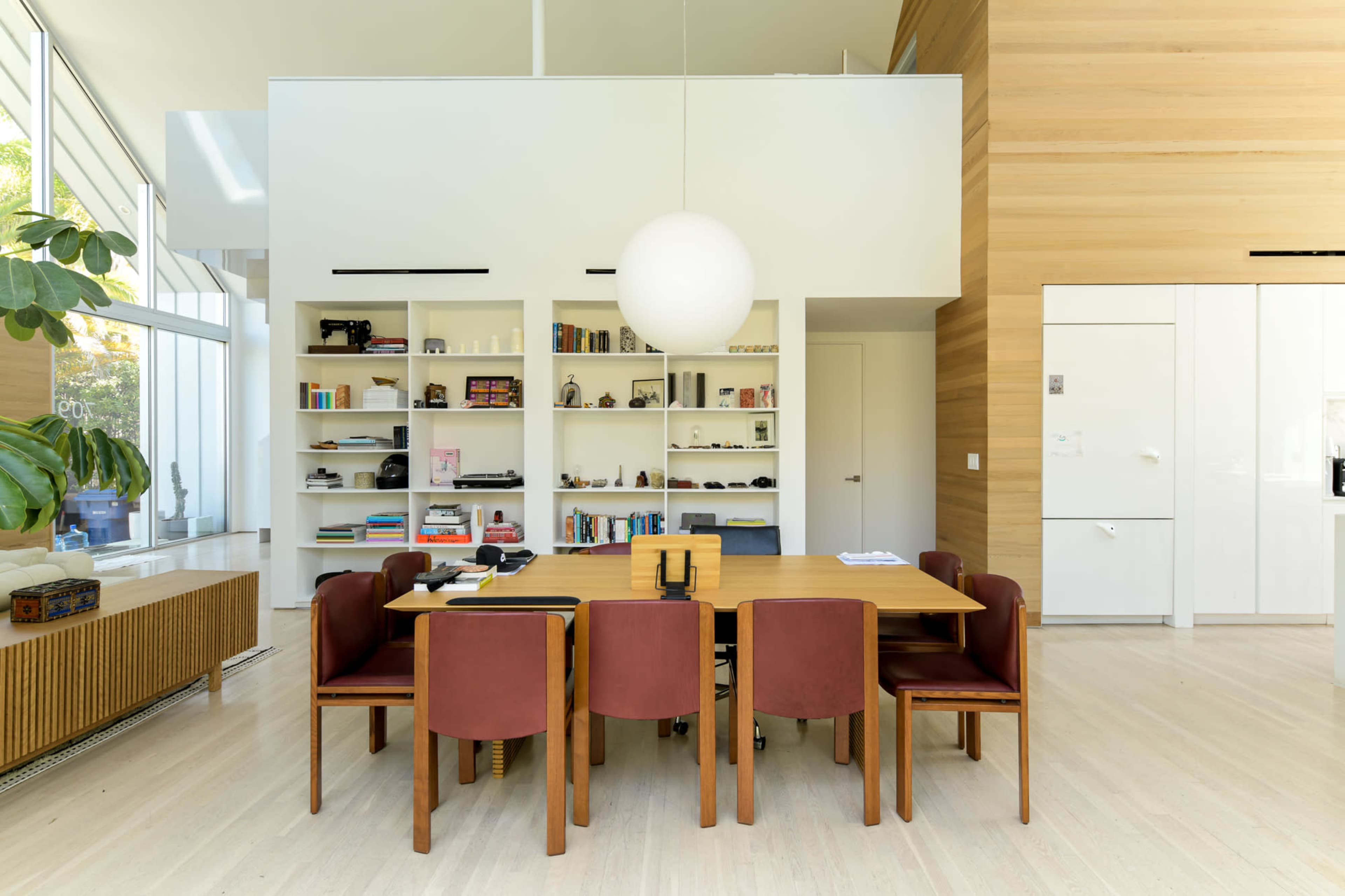 A modern dining area with a wooden table surrounded by six brown chairs, bookshelves in the background, and a large spherical light fixture hanging from the ceiling.