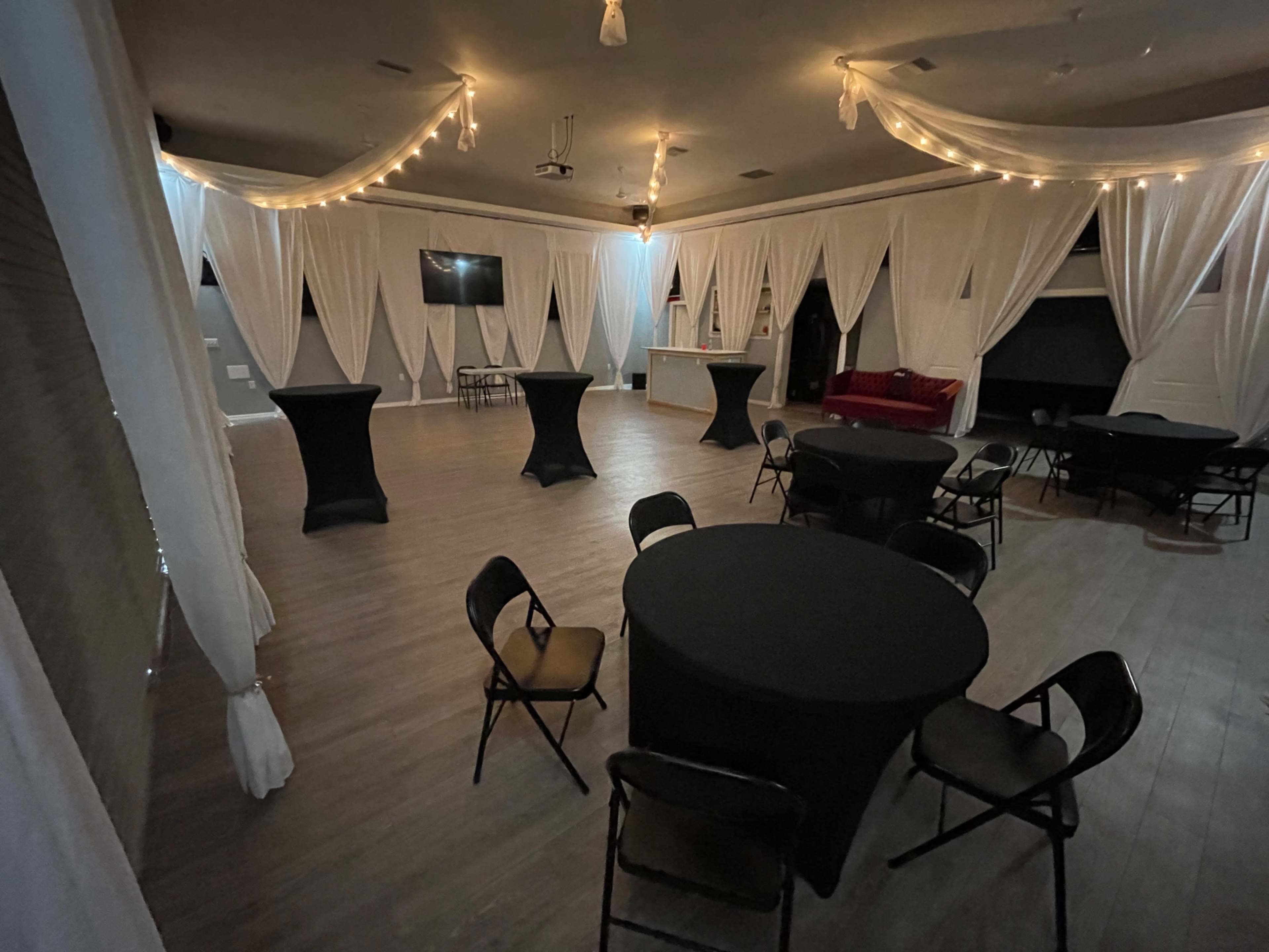 The image shows an event space with several round tables covered in black tablecloths, draped white curtains, and lights hanging from the ceiling.
