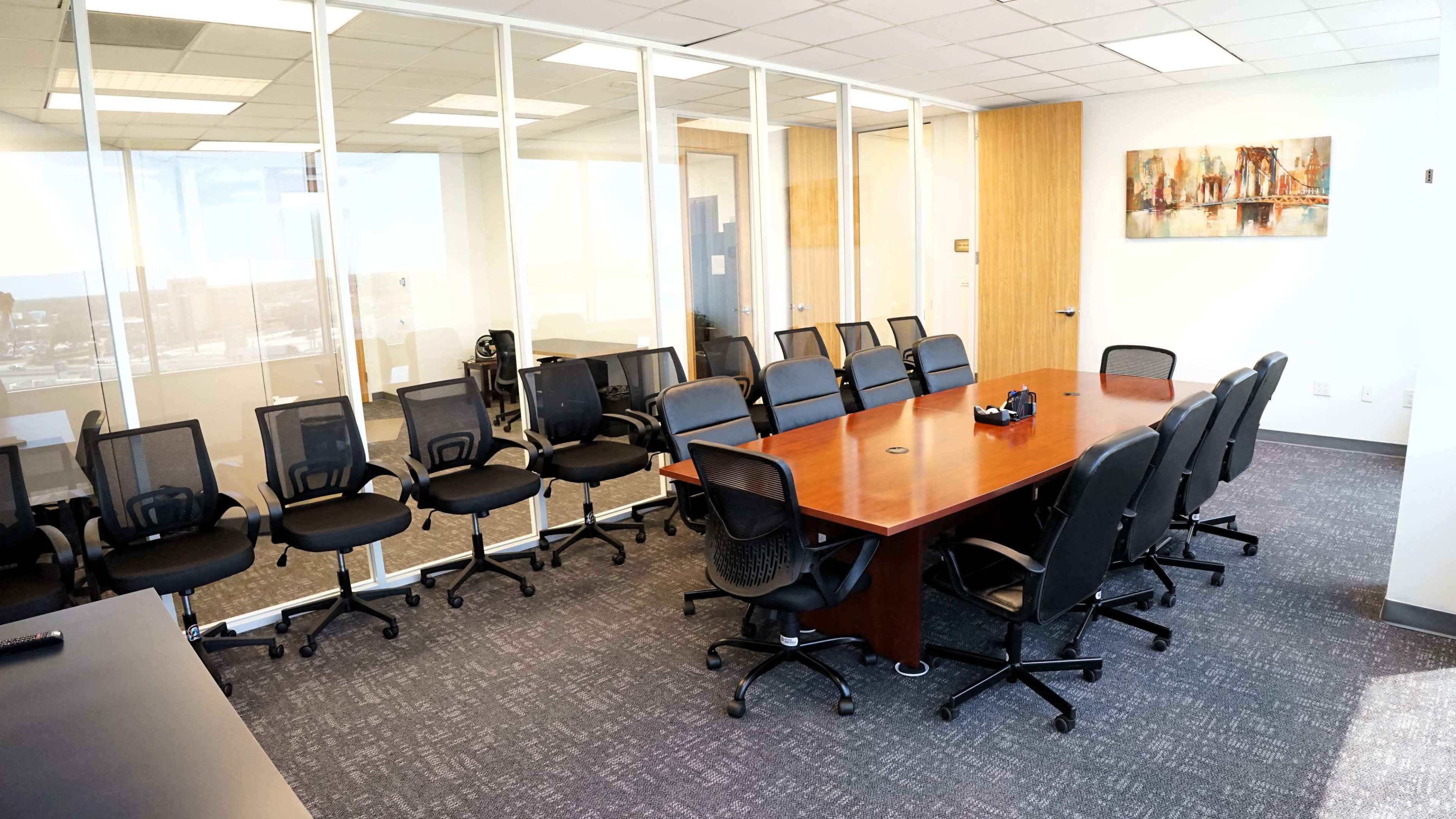 A modern conference room features a large wooden table surrounded by black office chairs and glass walls with a city view.