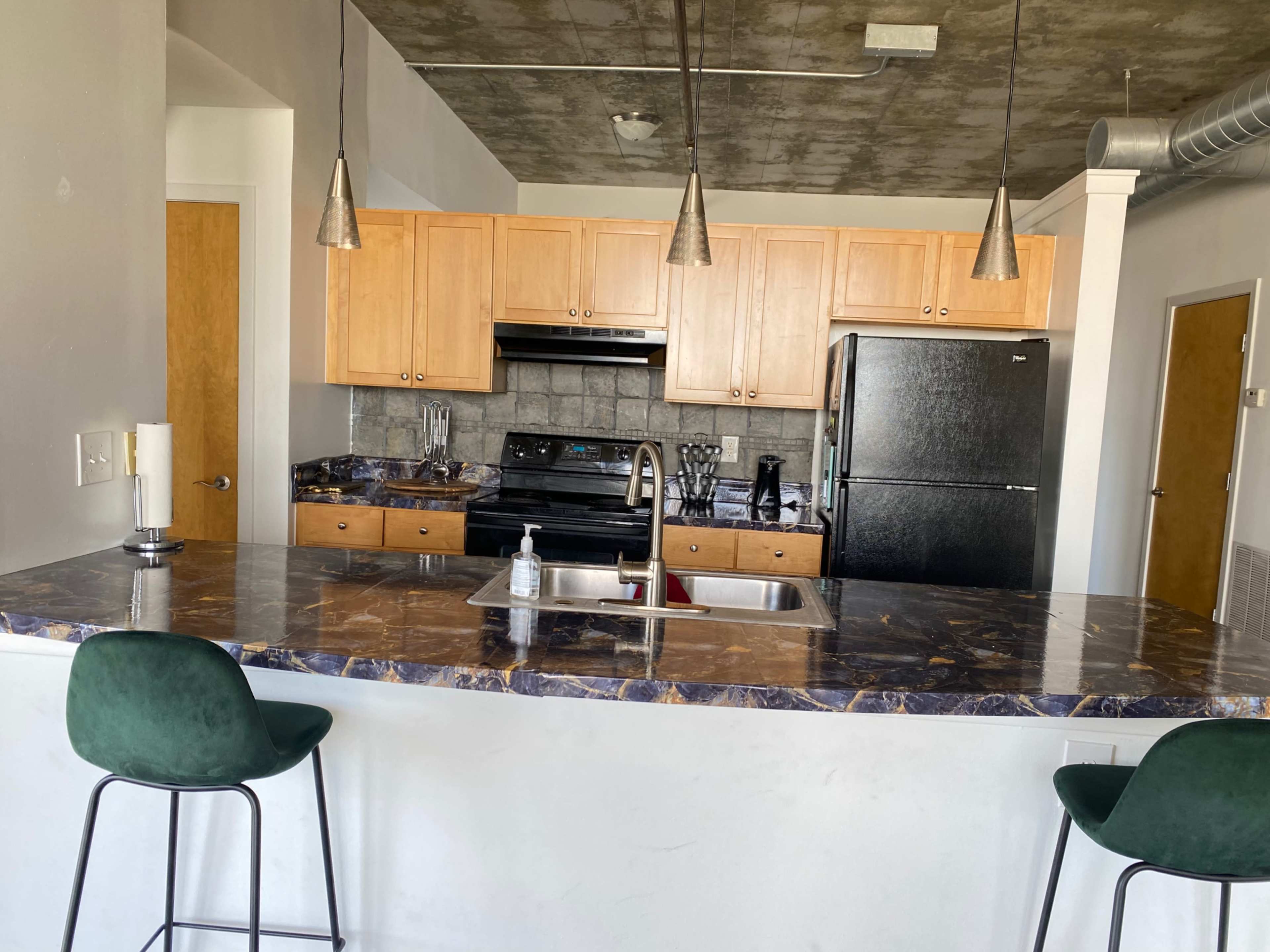 A modern kitchen with wooden cabinets, a black refrigerator, and a marble countertop with two green bar stools.