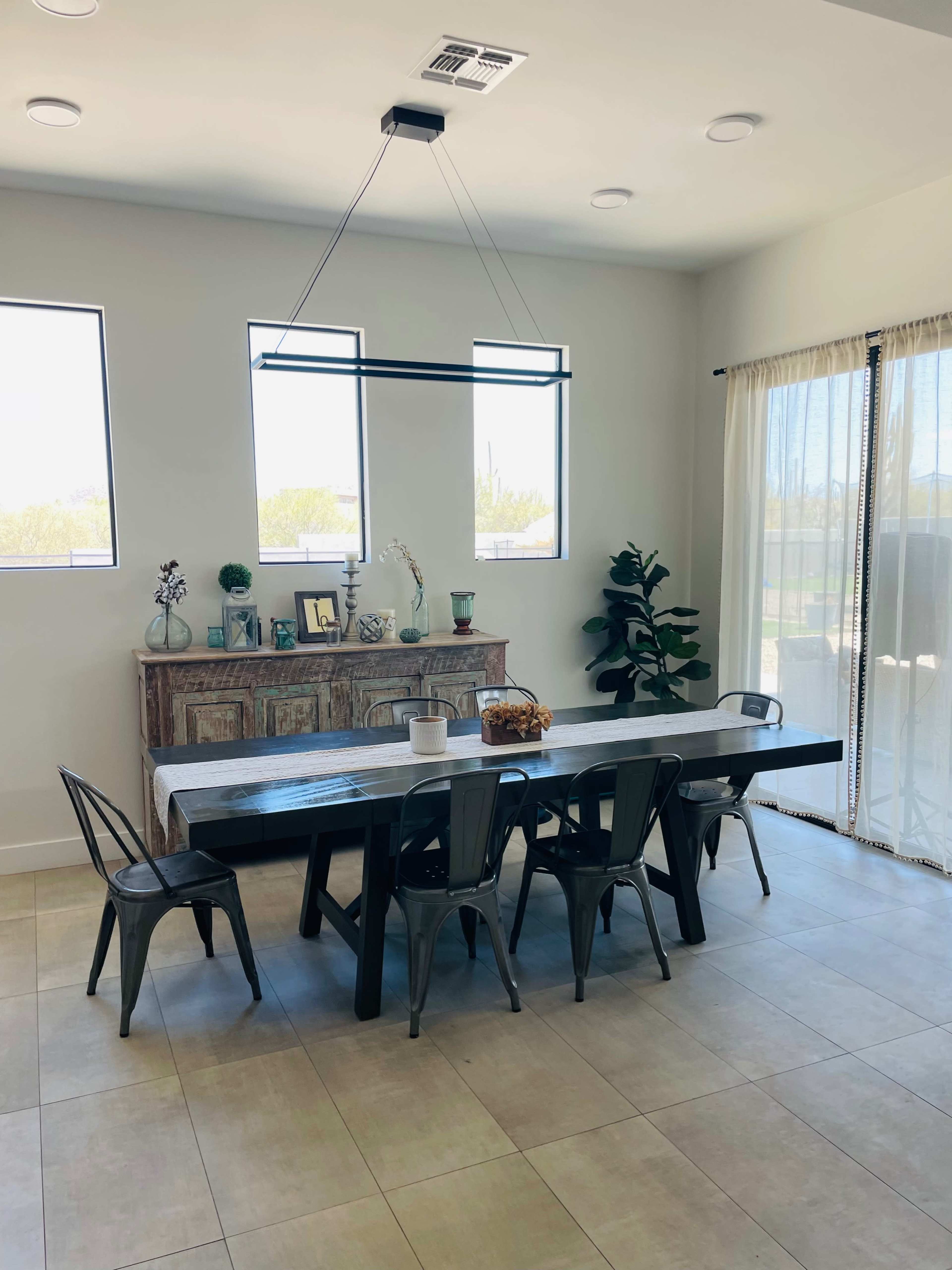A spacious dining area features a long table surrounded by metal chairs, with a sideboard and plants against the wall, illuminated by natural light from large windows.