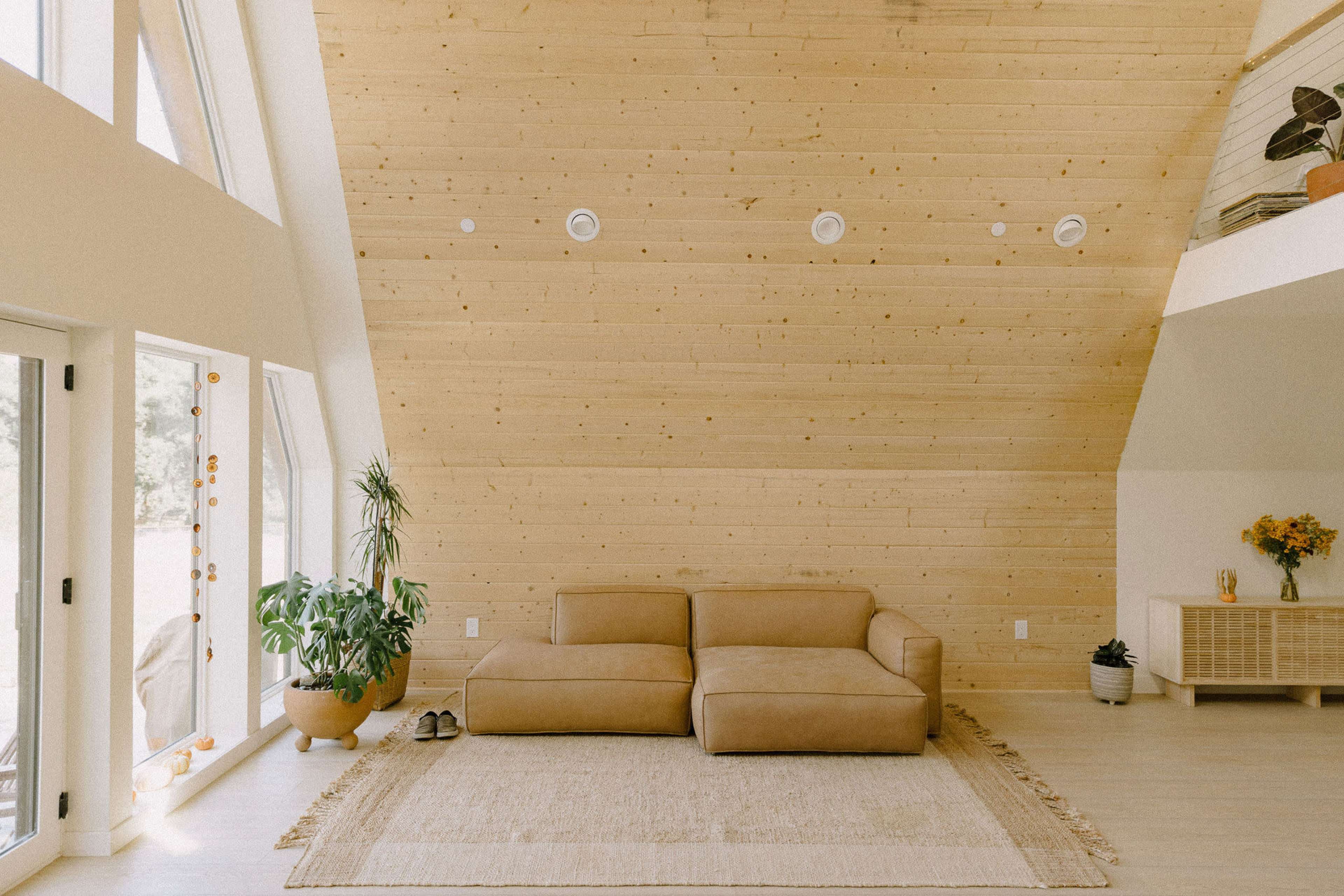 The image features a modern living room with a sloped wooden ceiling, a tan sectional sofa, and large windows that invite natural light.