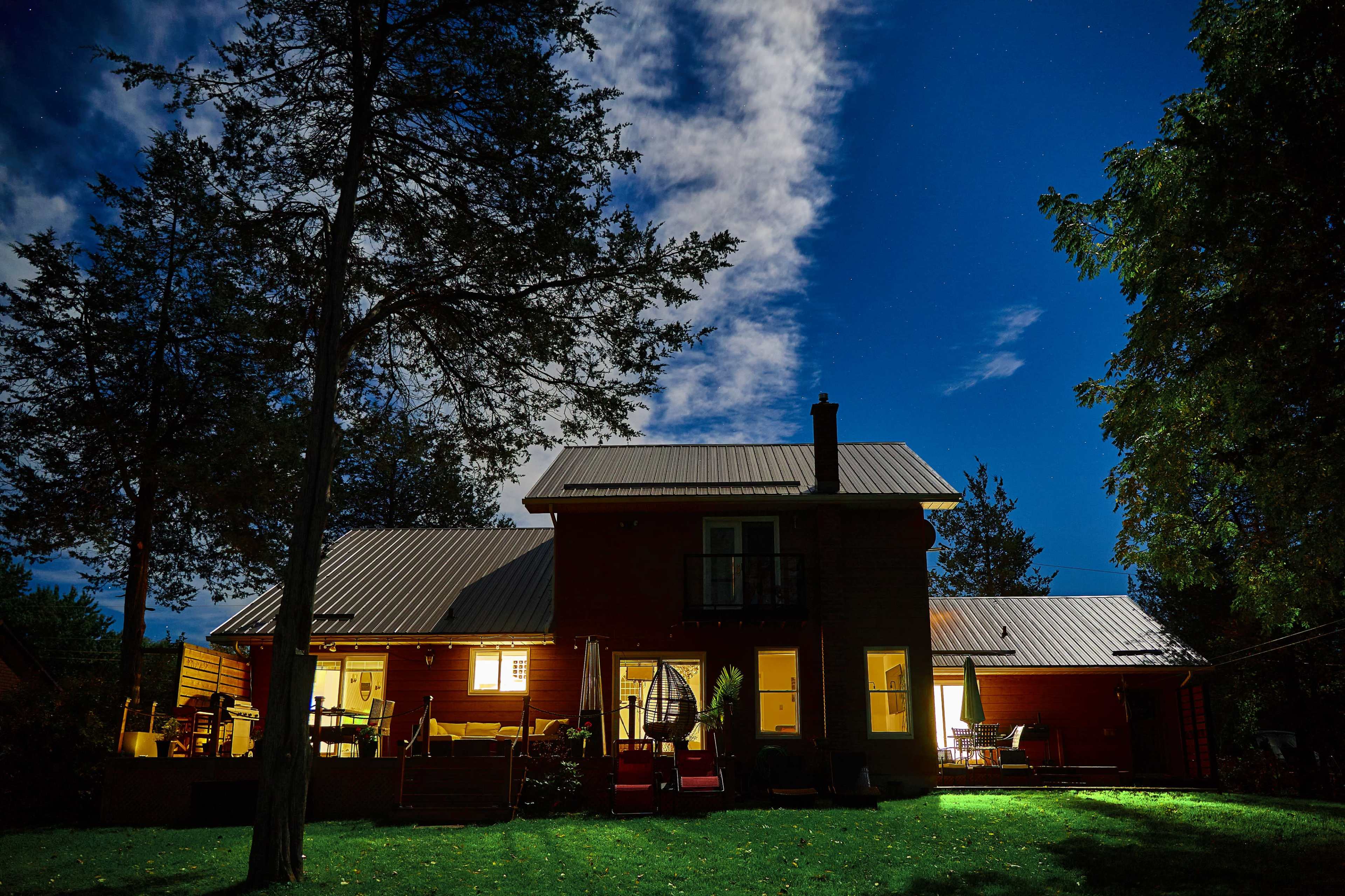 A red house with a metal roof is illuminated at night, surrounded by trees and a clear sky dotted with stars.