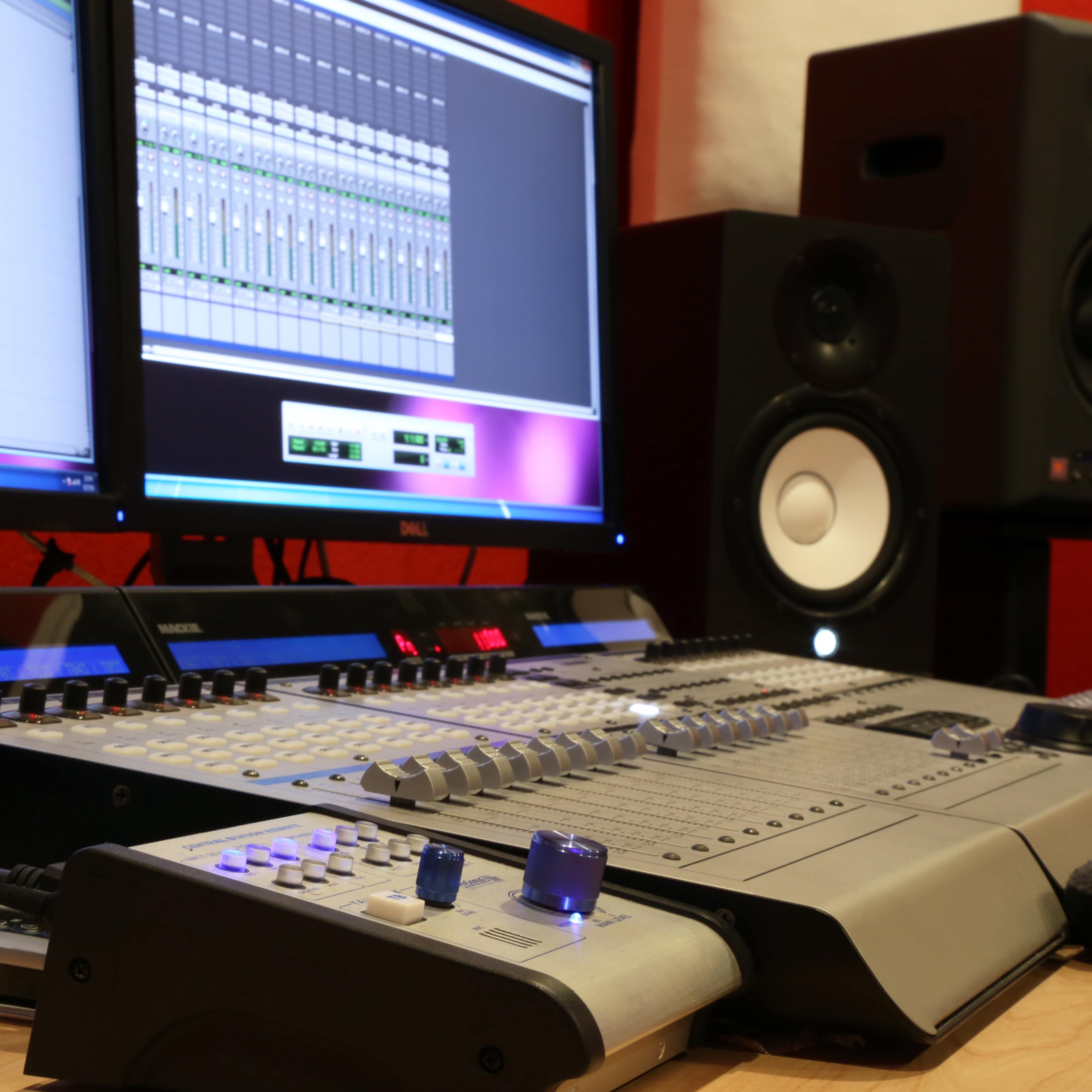 A digital audio mixing console in a sound studio, with a computer monitor displaying audio tracks and two studio speakers in the background.