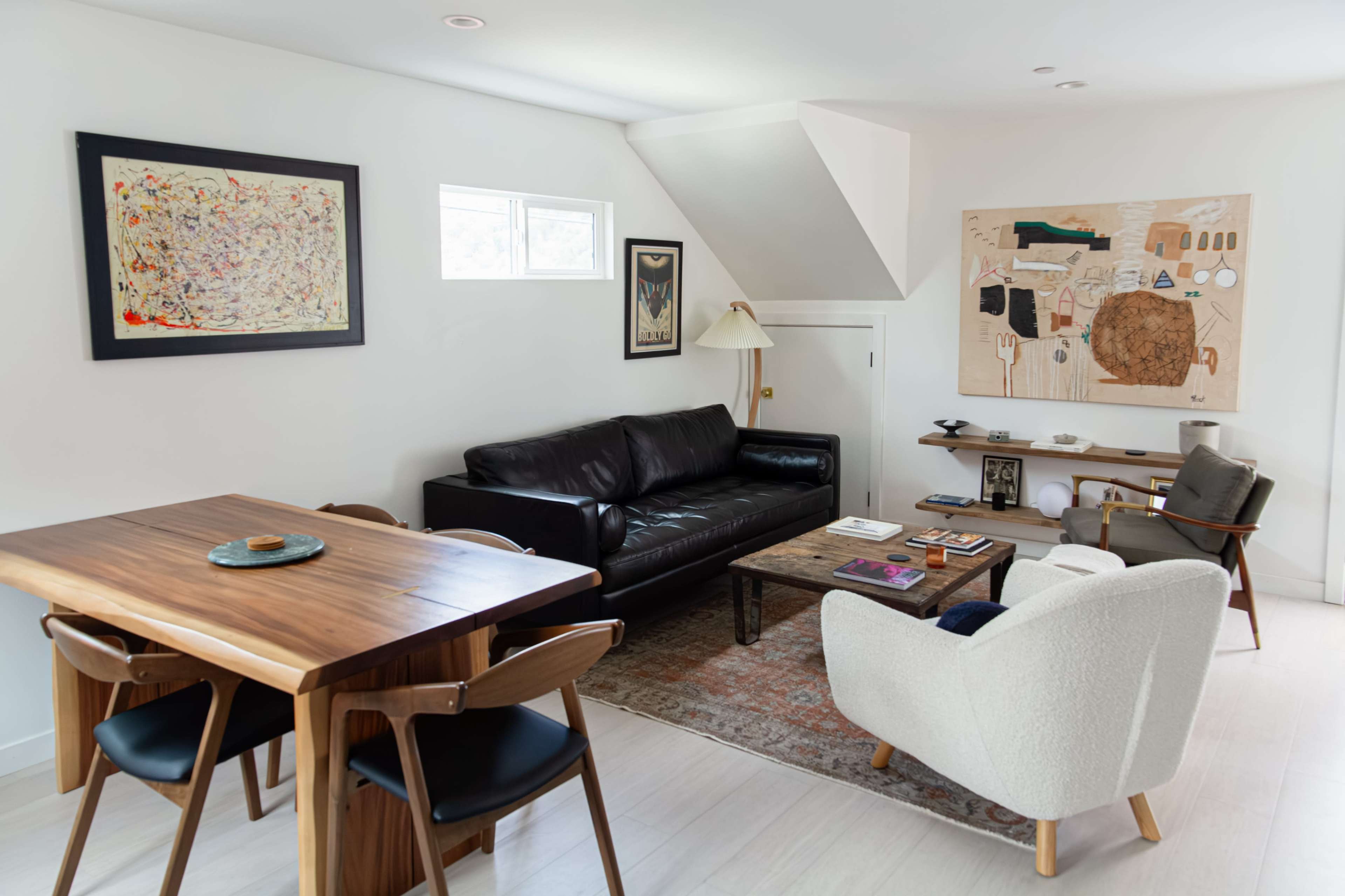 The image shows a modern living room featuring a black leather sofa, a wooden dining table with chairs, and decorative art on the white walls.
