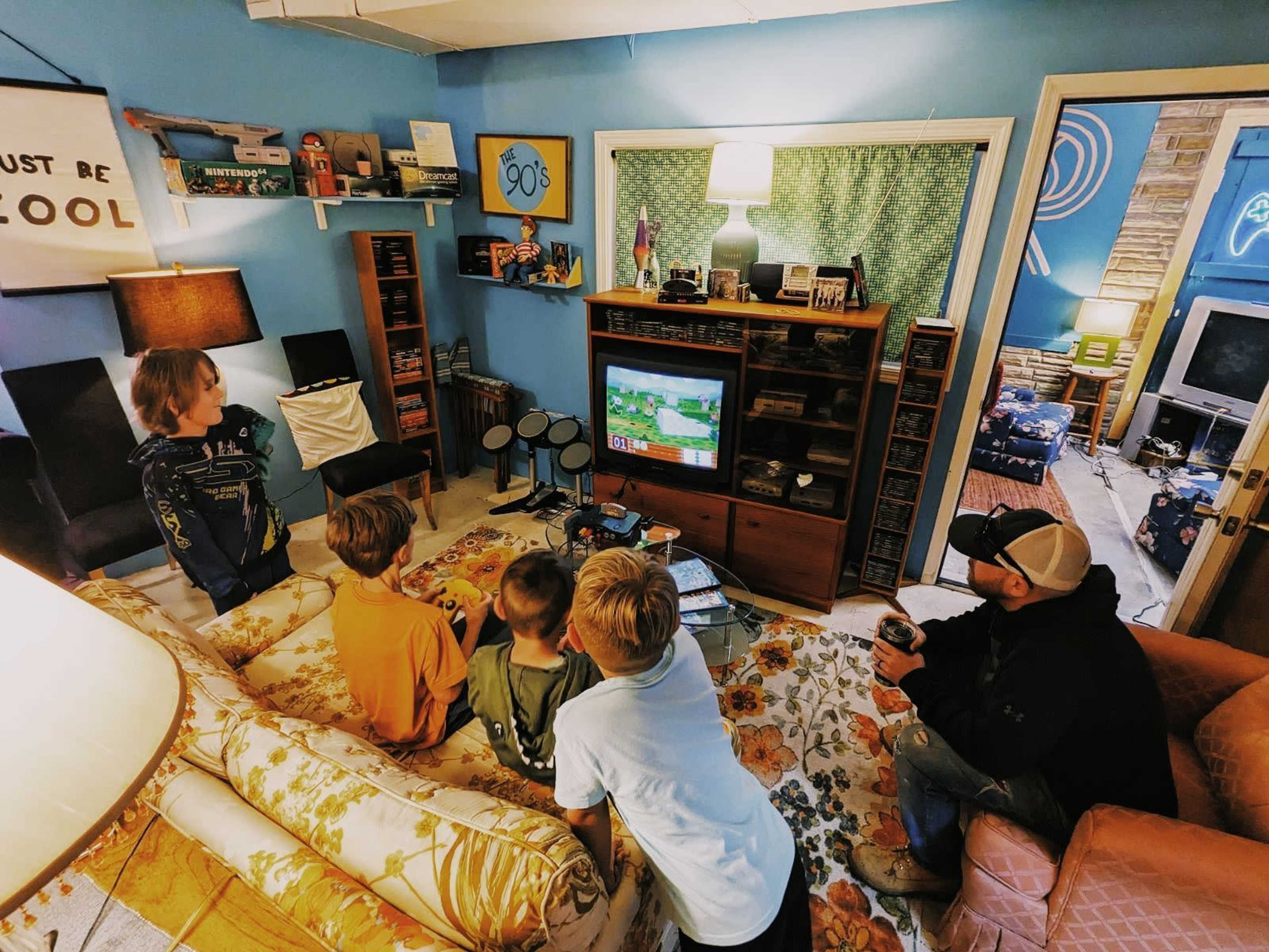 A group of children and an adult sit in a retro-style living room, watching a video game on a television.