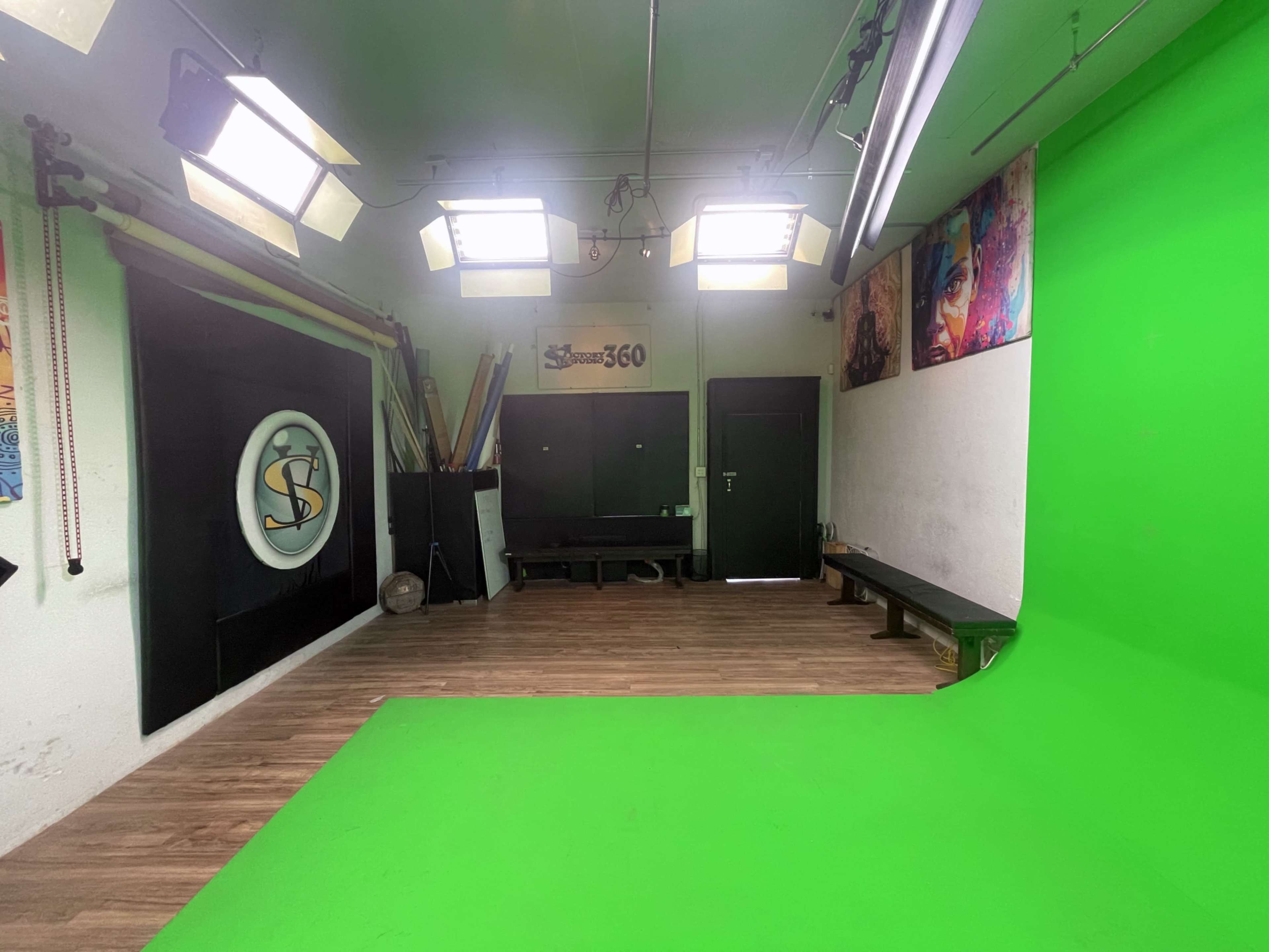 The image shows a studio with a green backdrop, multiple bright lights, and a plain wall adorned with artwork.