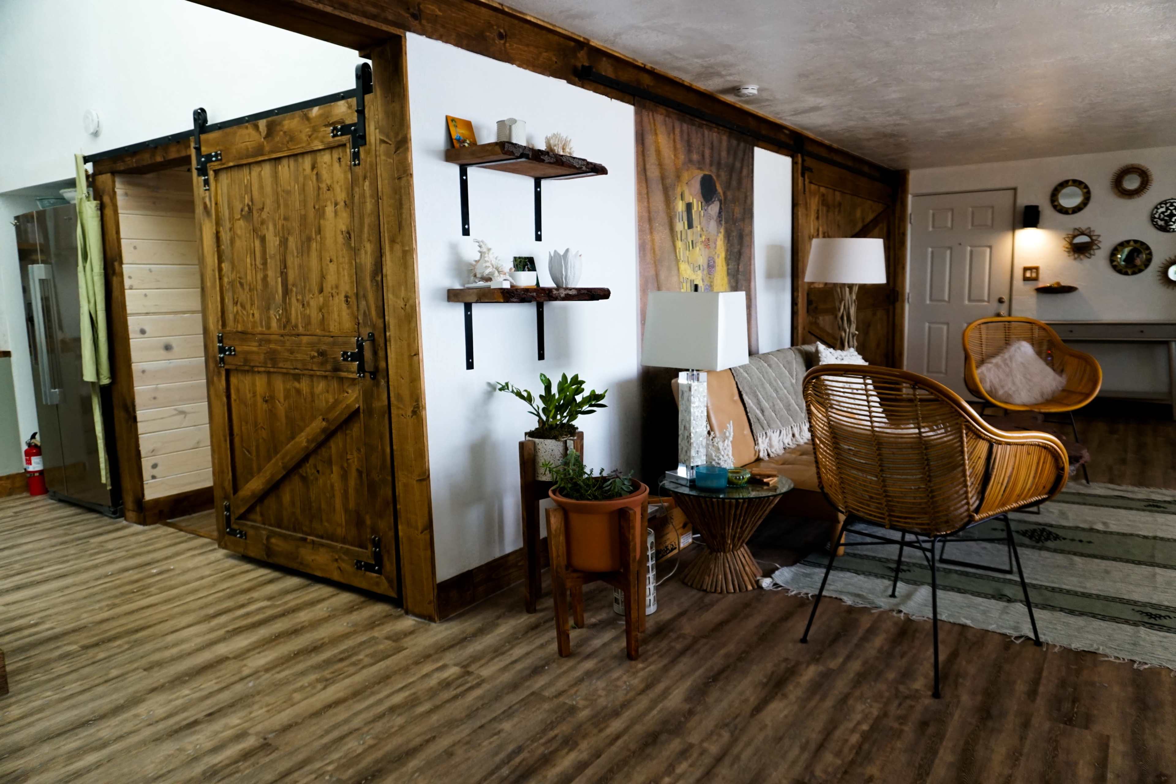 The image shows a rustic living room with wooden accents, a barn-style sliding door, a cozy seating area, and shelves displaying decor items.