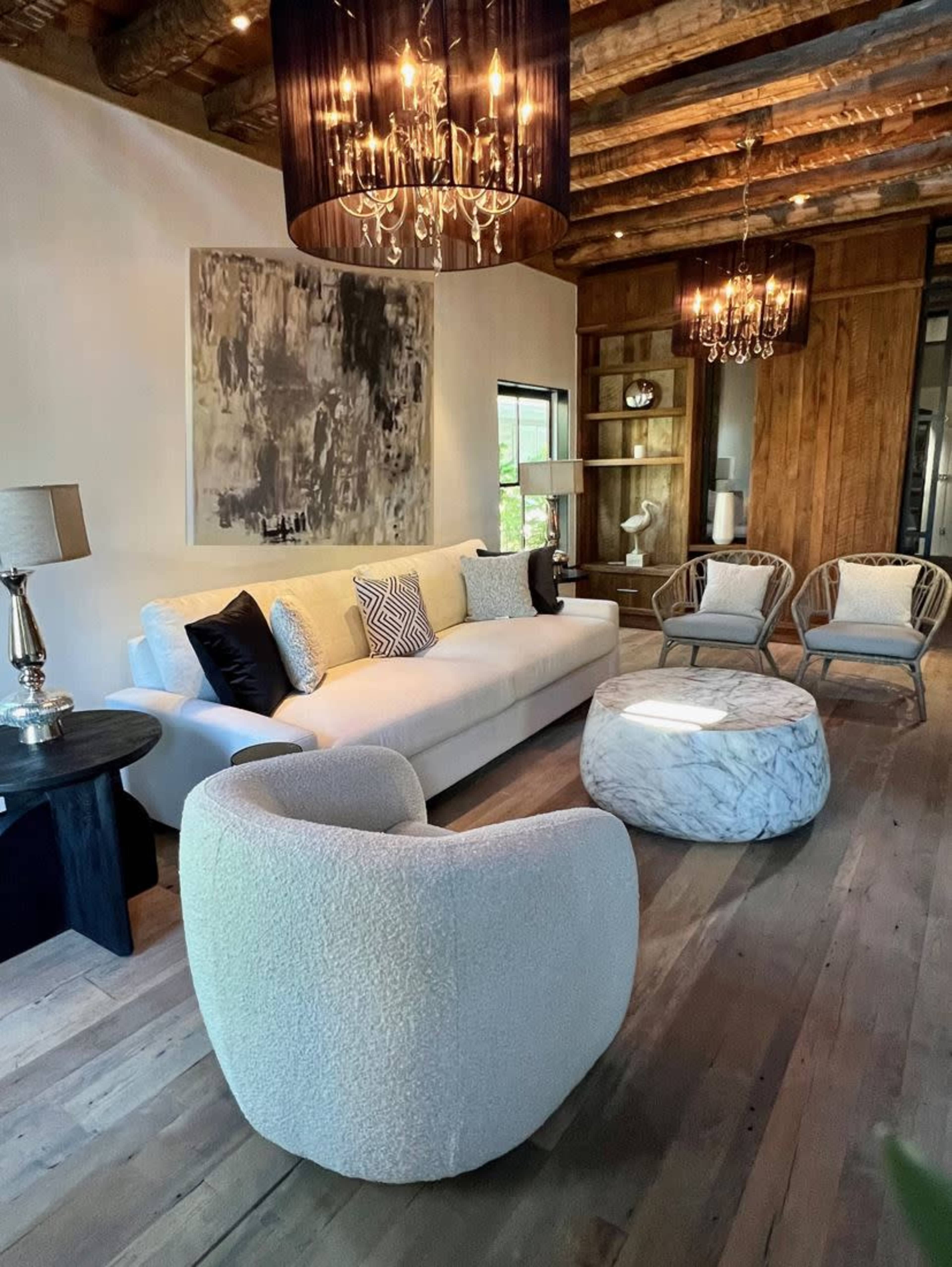 The room features a modern seating area with a white sofa, two chairs, and a round, textured ottoman under a ceiling with wooden beams and multiple chandeliers.