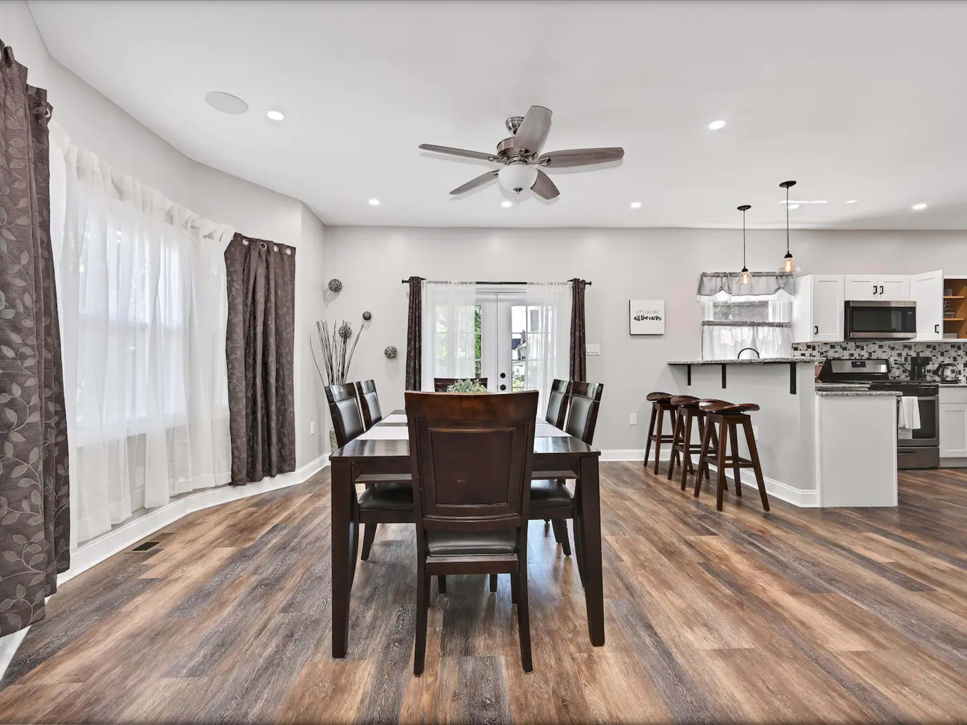 The image shows a dining area with a wooden table and chairs, surrounded by large windows and adjacent to a modern kitchen.