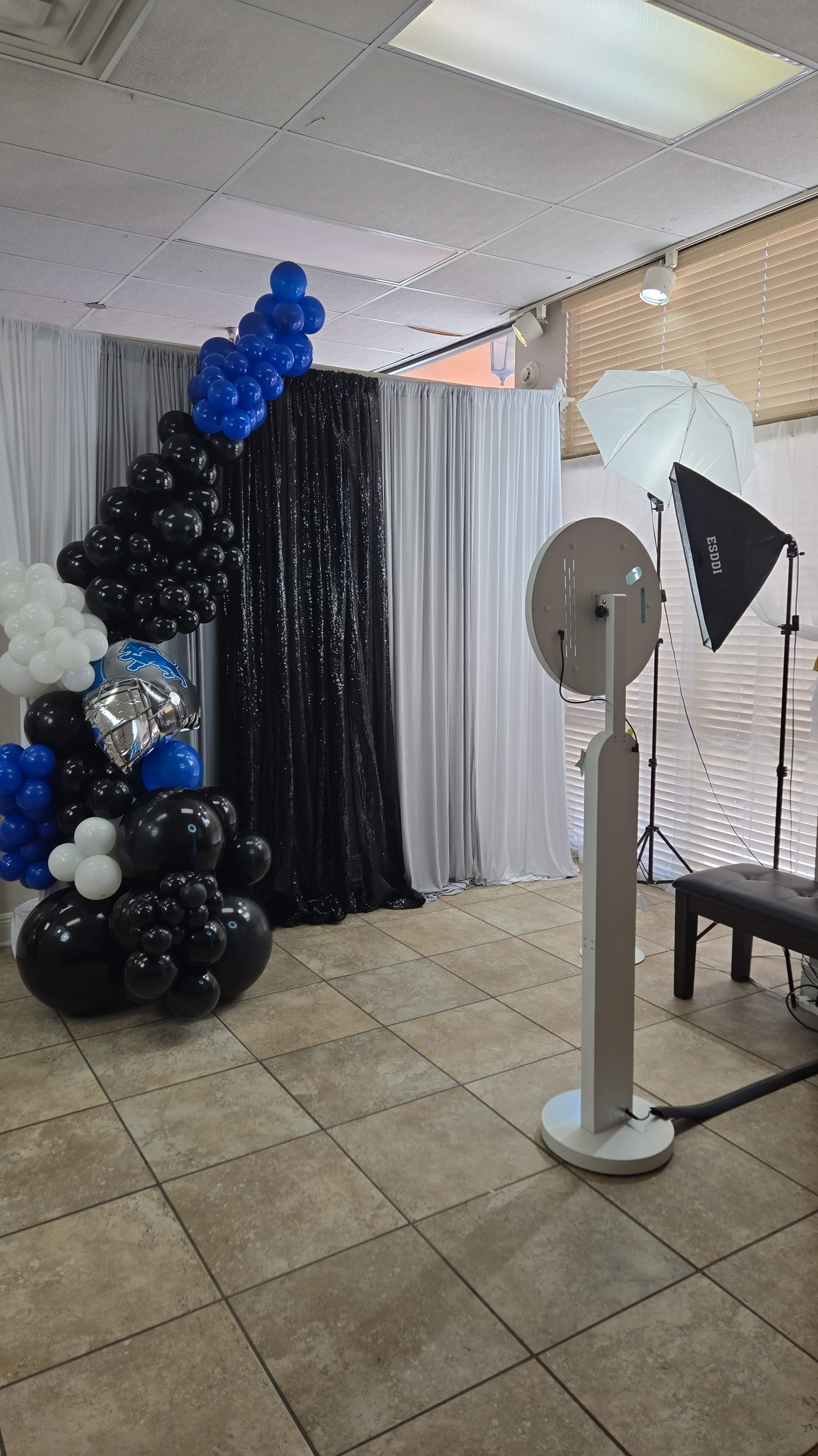 The image shows a decorated photo booth area featuring black, white, and blue balloons, a backdrop curtain, and photography equipment including a softbox light and a camera.