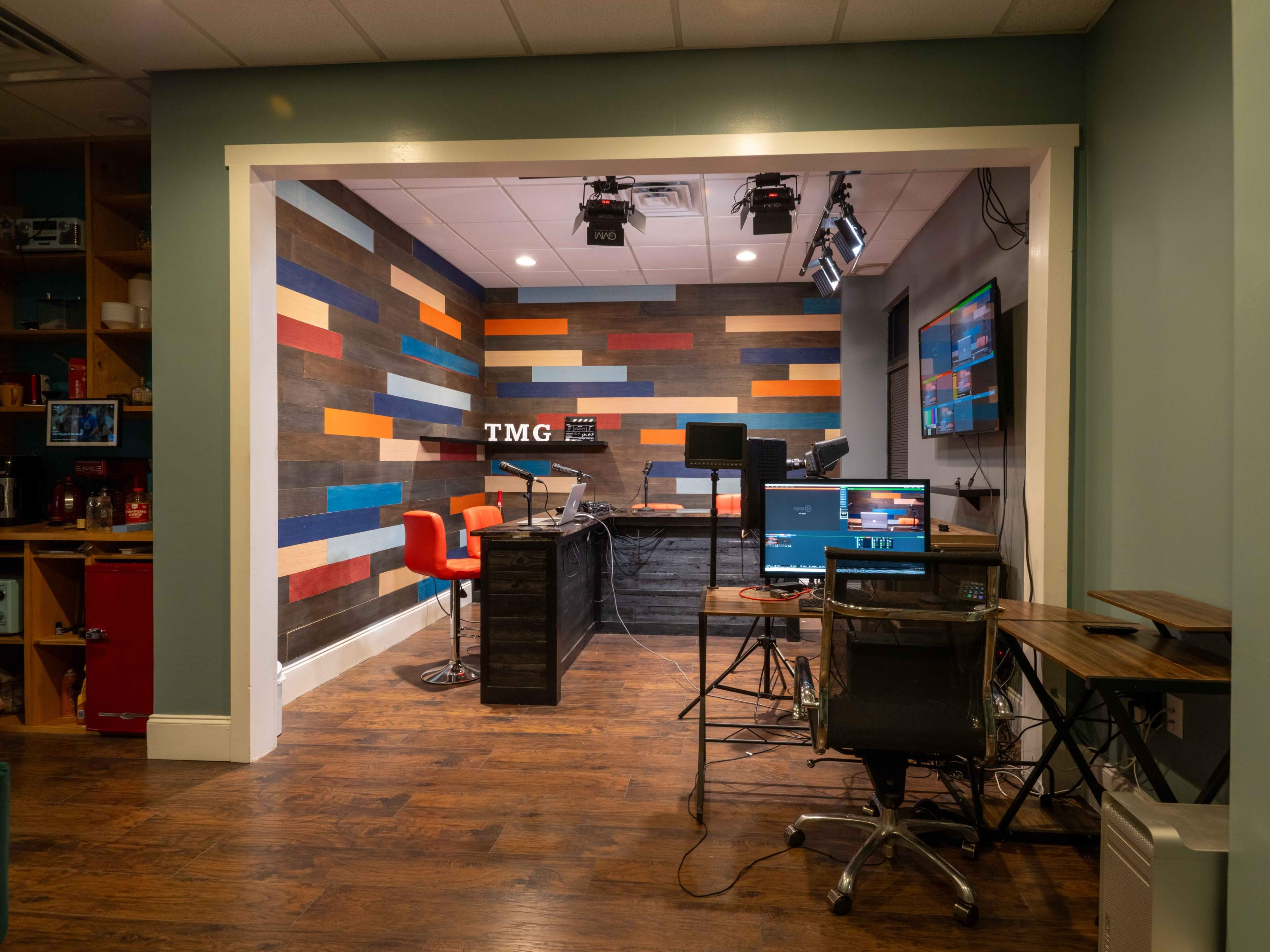 A modern broadcast studio featuring a desk with multiple screens, colorful wall panels, and an orange chair.