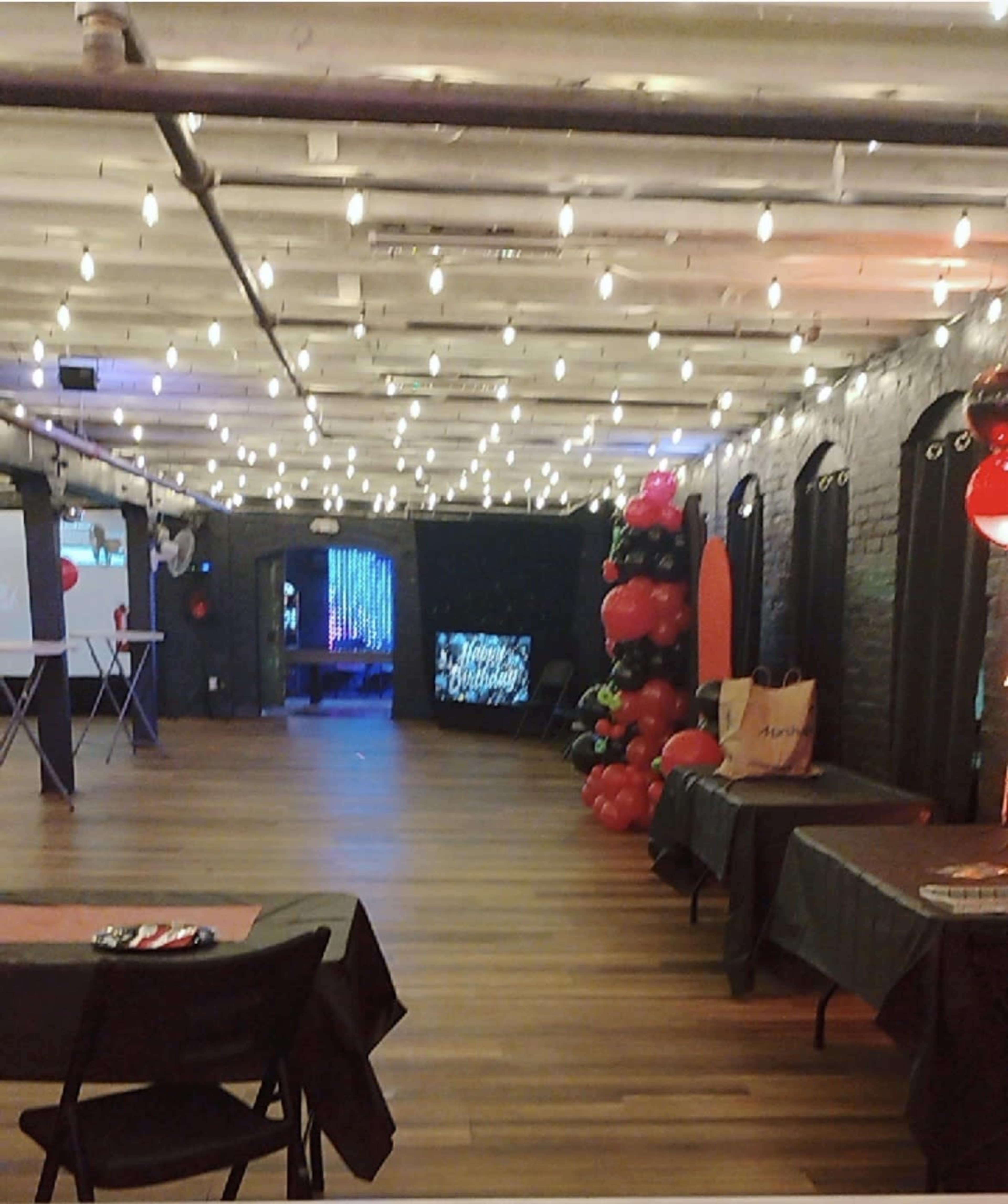 The image shows an indoor event space with wooden floors, string lights overhead, and decorated tables along the sides.