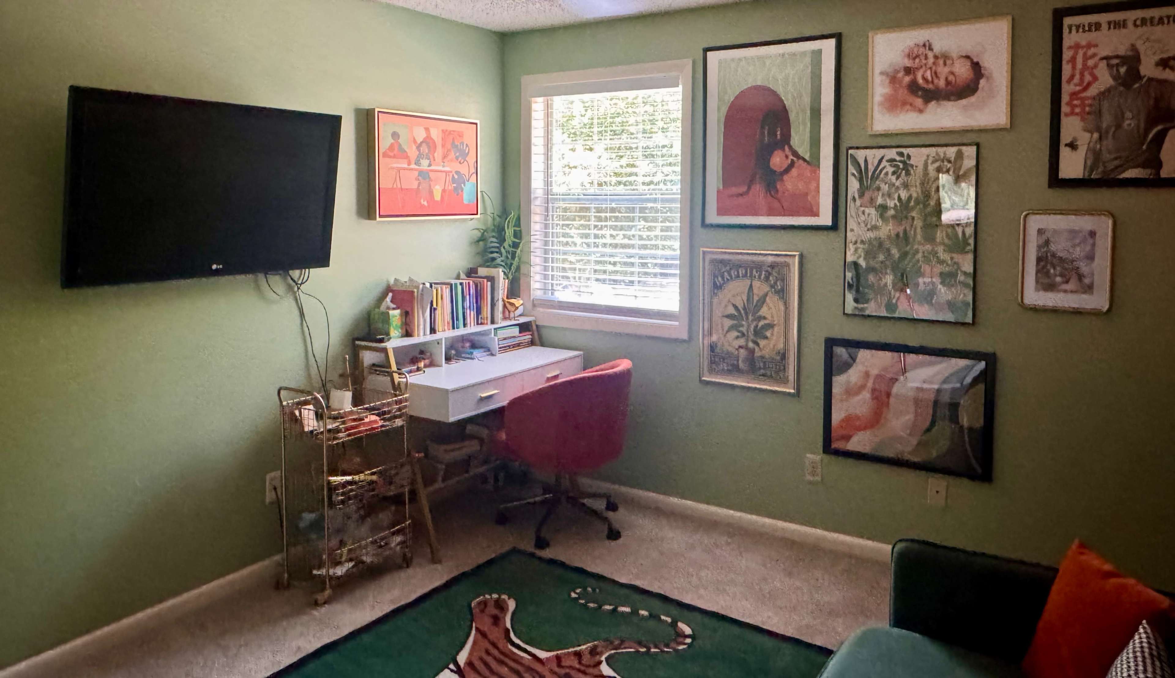 The room features a green wall, a mounted TV, a desk with a red chair, shelves with books, and various framed artworks displayed on the walls.
