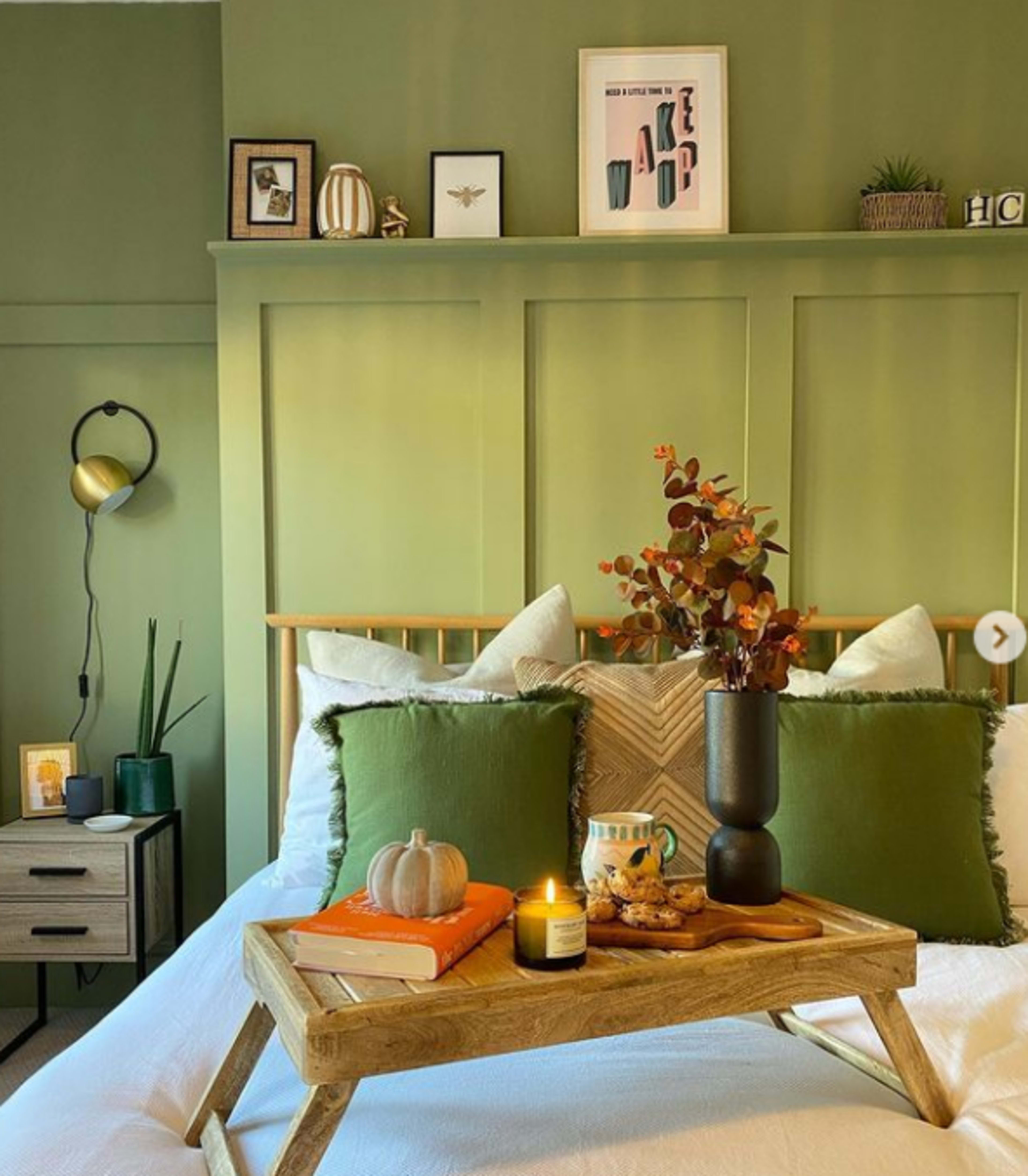 A neatly arranged bedroom features a wooden bed with green and white pillows, a decorative tray with a book, candle, and snacks, alongside a nightstand and wall art in shades of green.