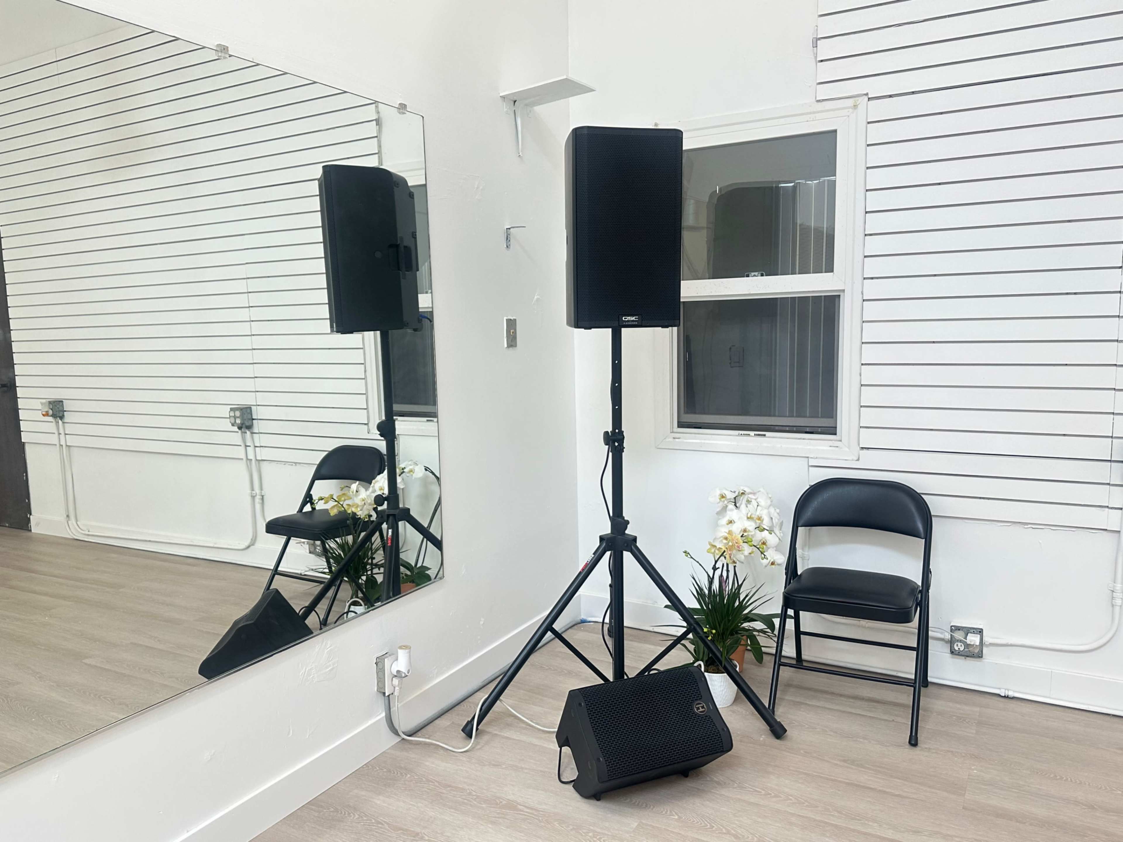 The image shows a simple indoor space with a black speaker on a stand, a folding chair, and a large mirror reflecting part of the room.