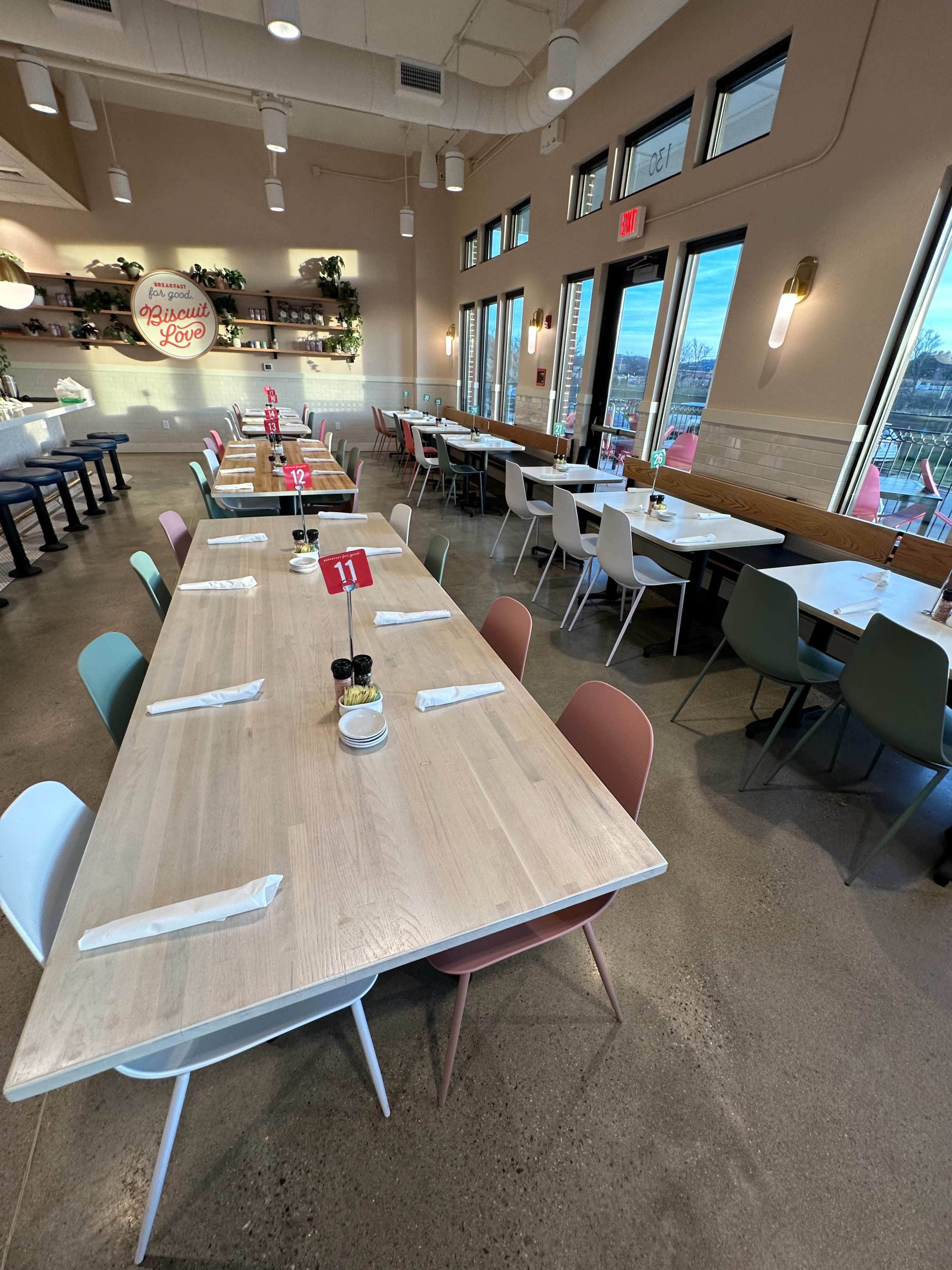 The image shows a modern restaurant interior featuring several tables arranged with place settings, surrounded by large windows that offer a view of the outdoors.