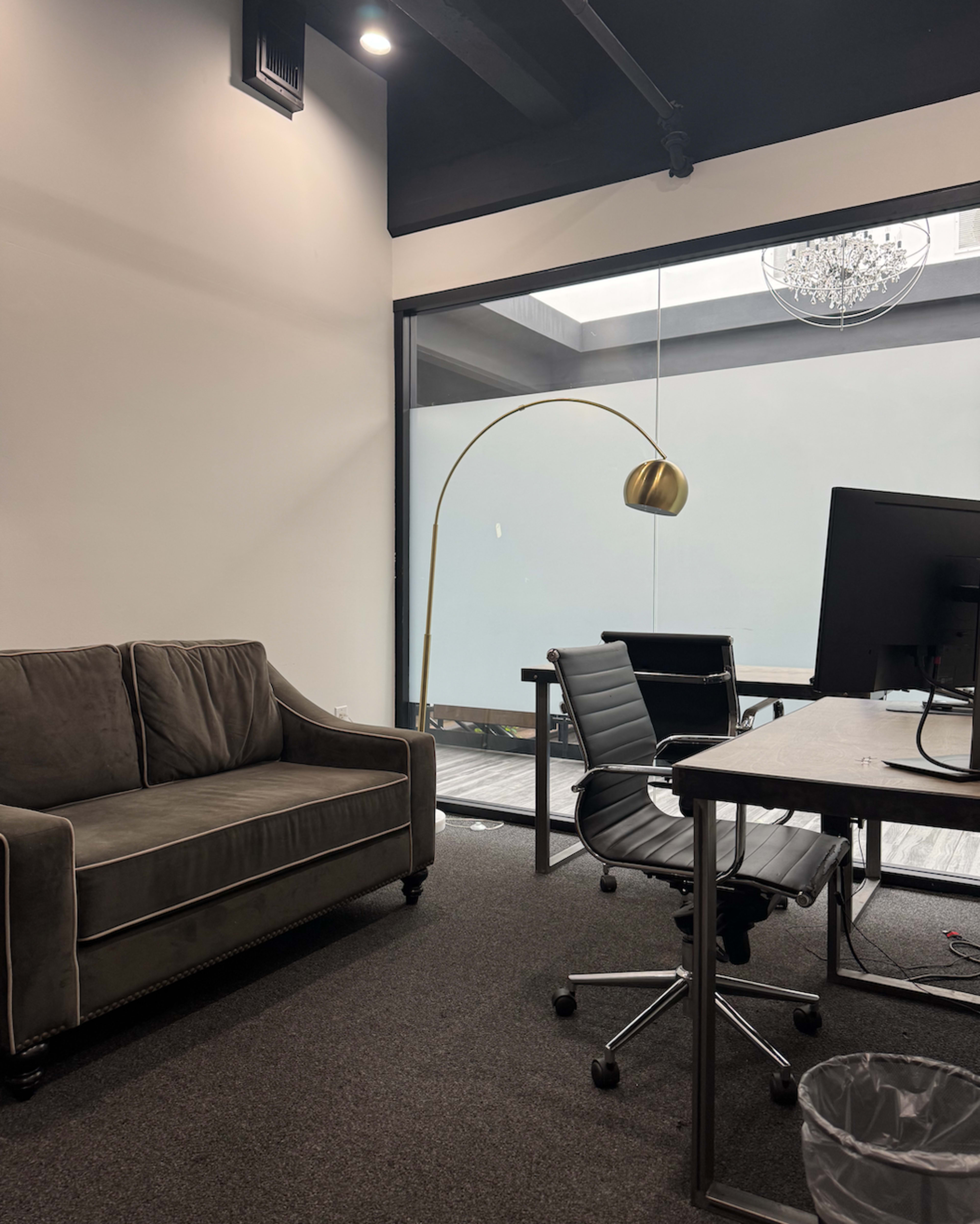 The image shows a modern office space with a gray sofa, a metal desk with a computer monitor, and a floor lamp, all set against a backdrop of a glass wall.