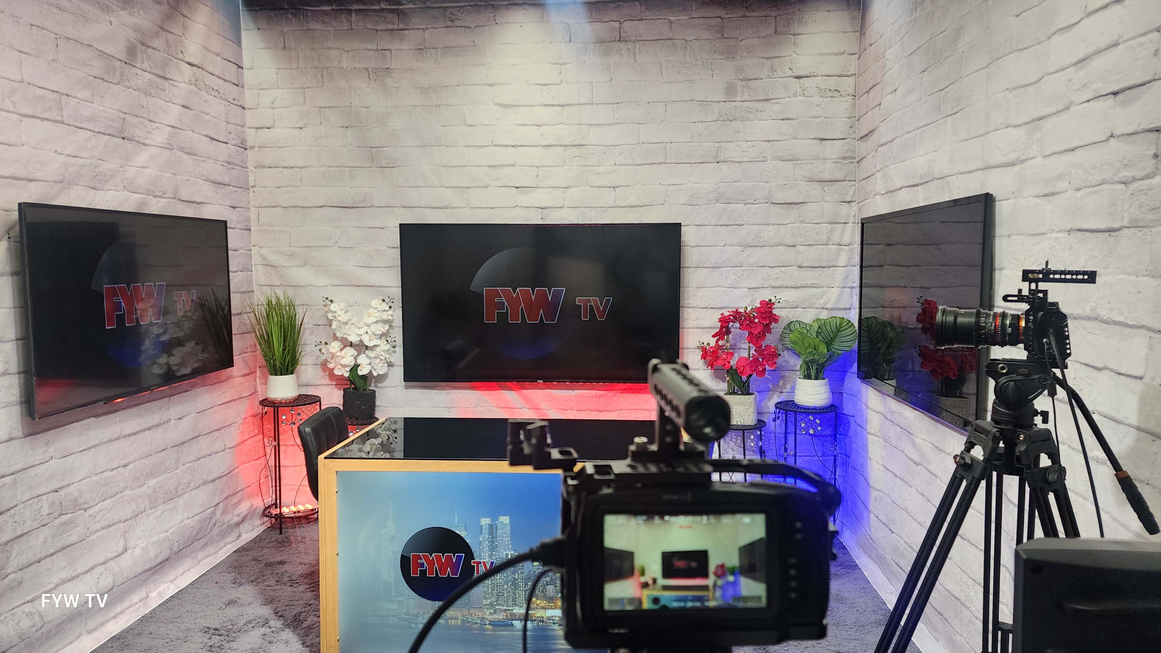 The image shows a television studio setup with multiple screens displaying the logo "FYW TV," surrounded by decorative plants and a backdrop of a brick wall.