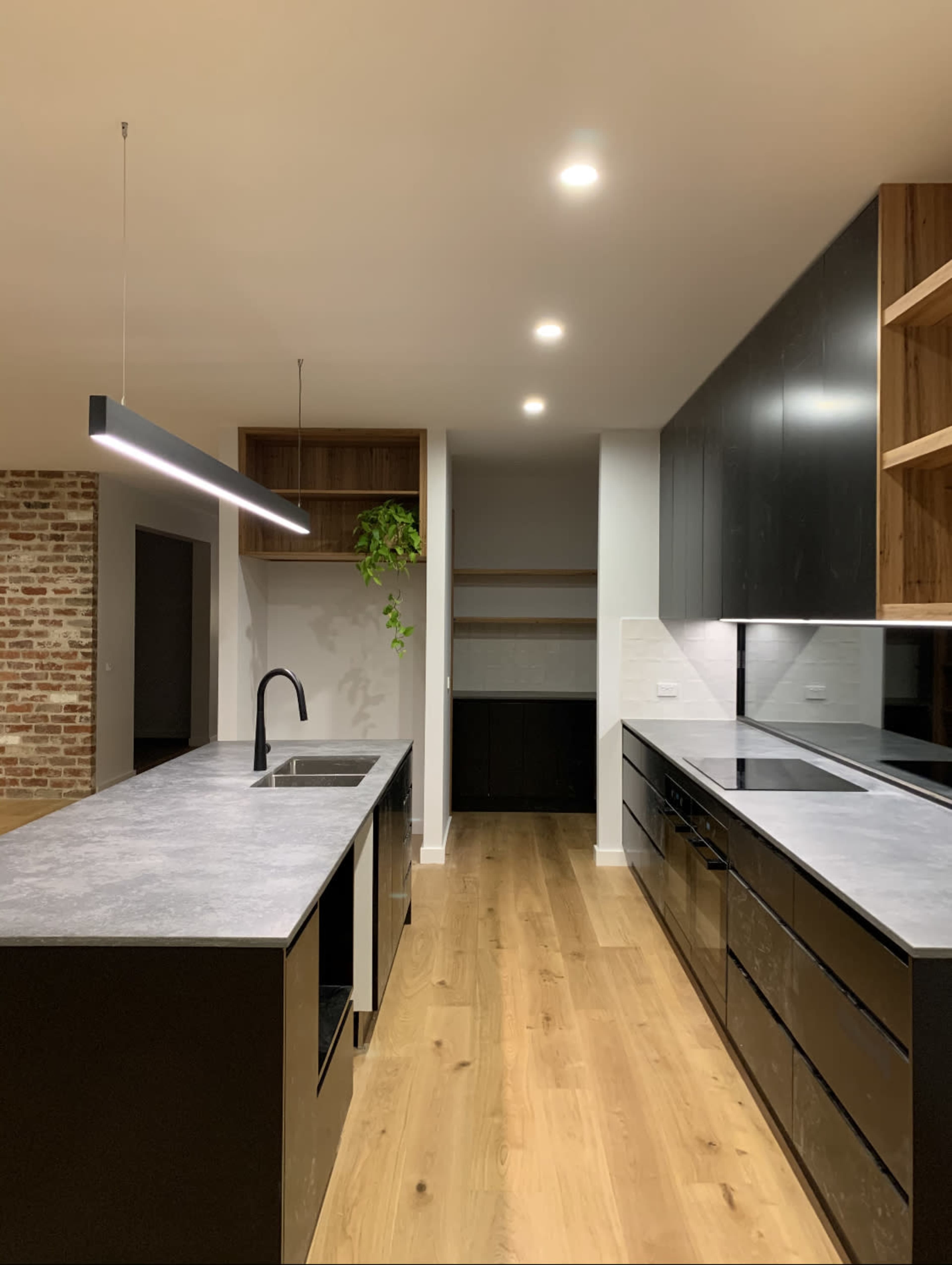 A modern kitchen with a central island featuring a sink, black cabinetry, wooden shelves, and an exposed brick wall.