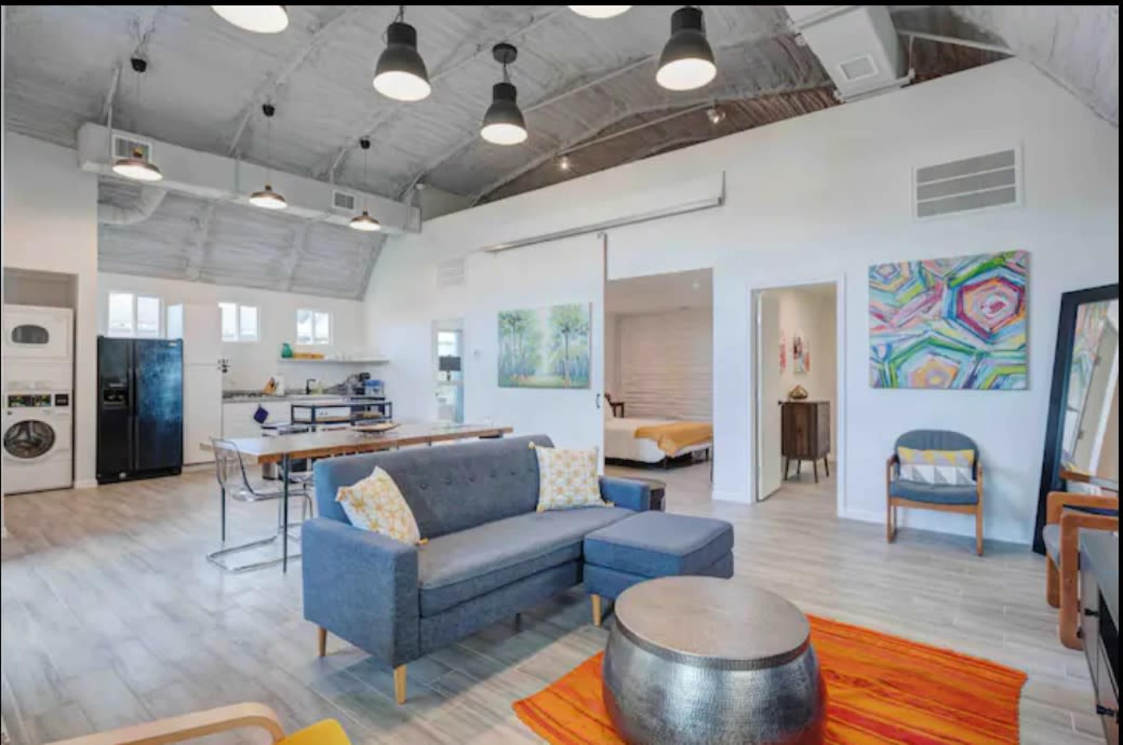 A modern, open-concept living space featuring a kitchen, dining area, and lounge with a blue sofa and colorful artwork on the walls.