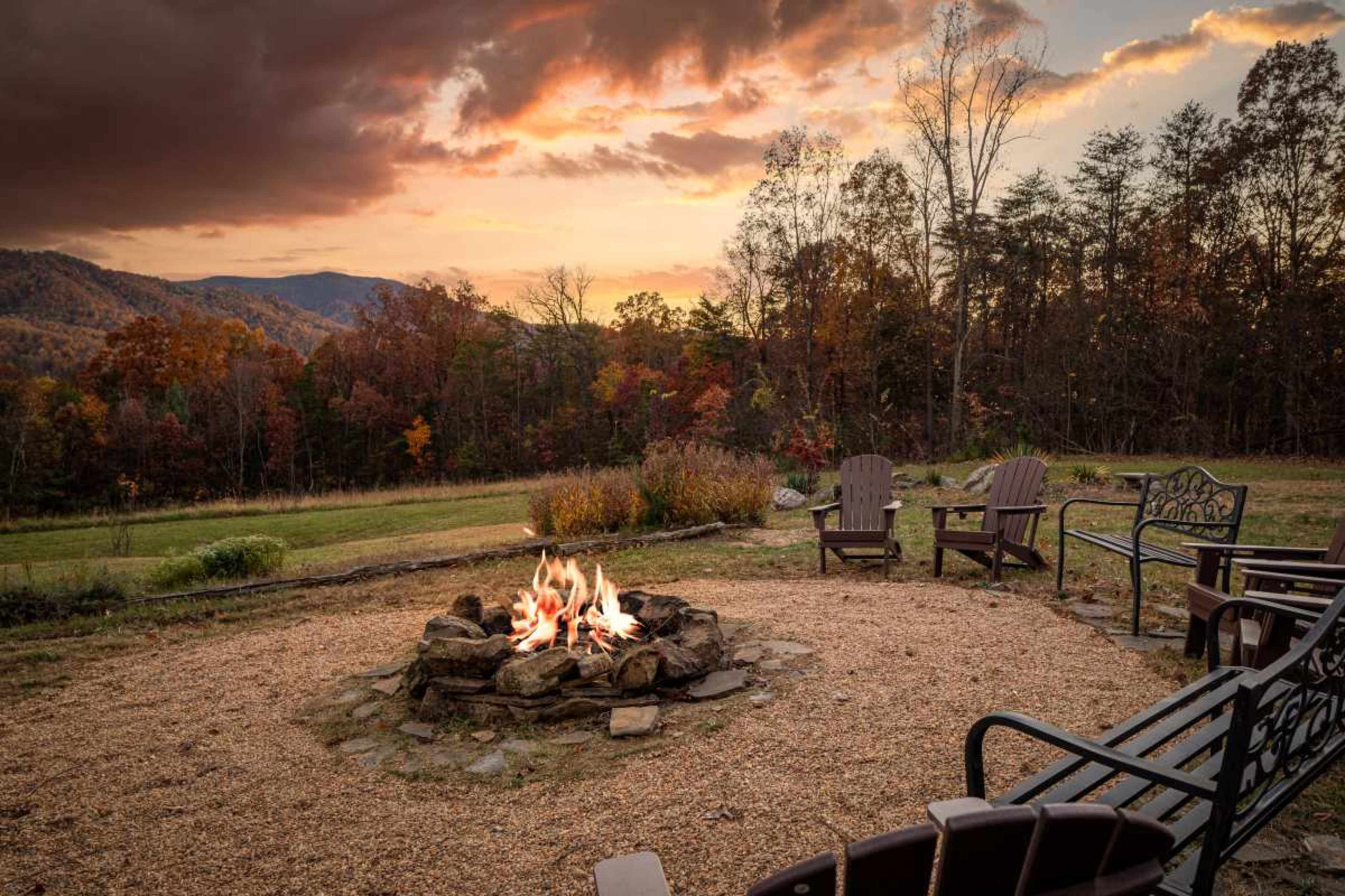 A campfire burns in a stone circle surrounded by chairs, with colorful autumn trees and mountains in the background under a cloudy sky.
