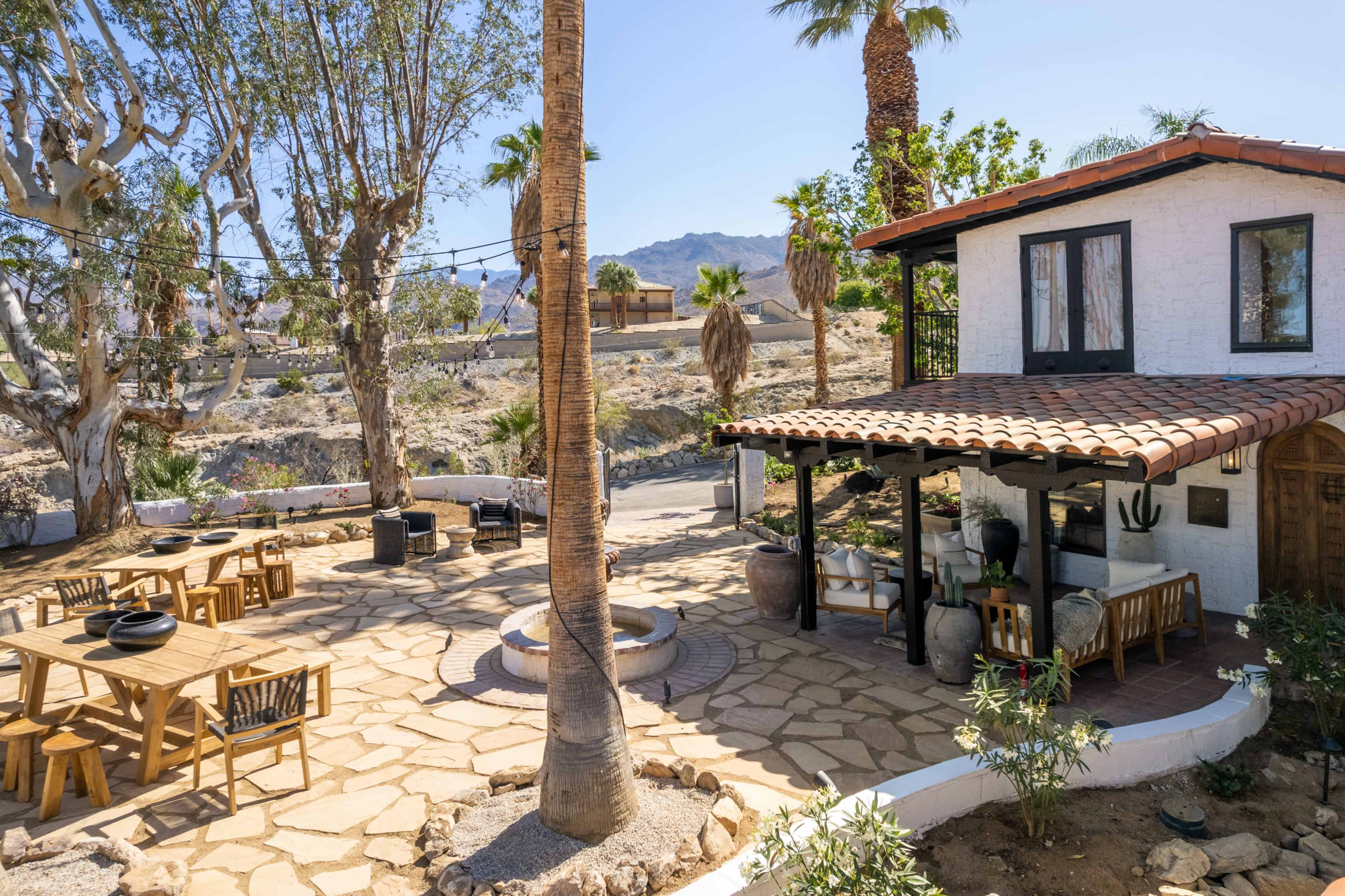 A house with a tiled roof and a stone patio is surrounded by palm trees and desert landscaping.