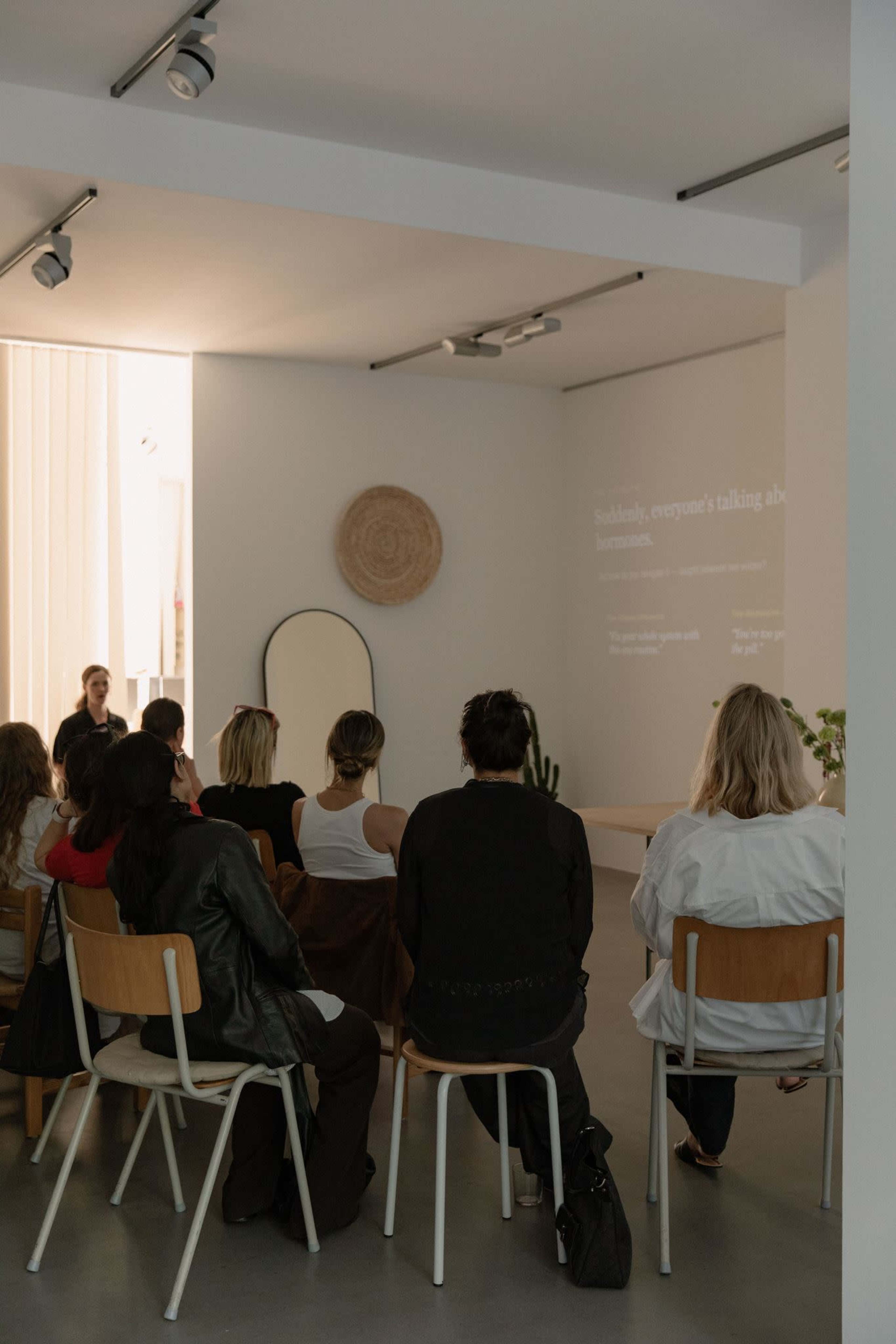 A group of people is seated in a bright room, listening to a presentation projected on a wall.