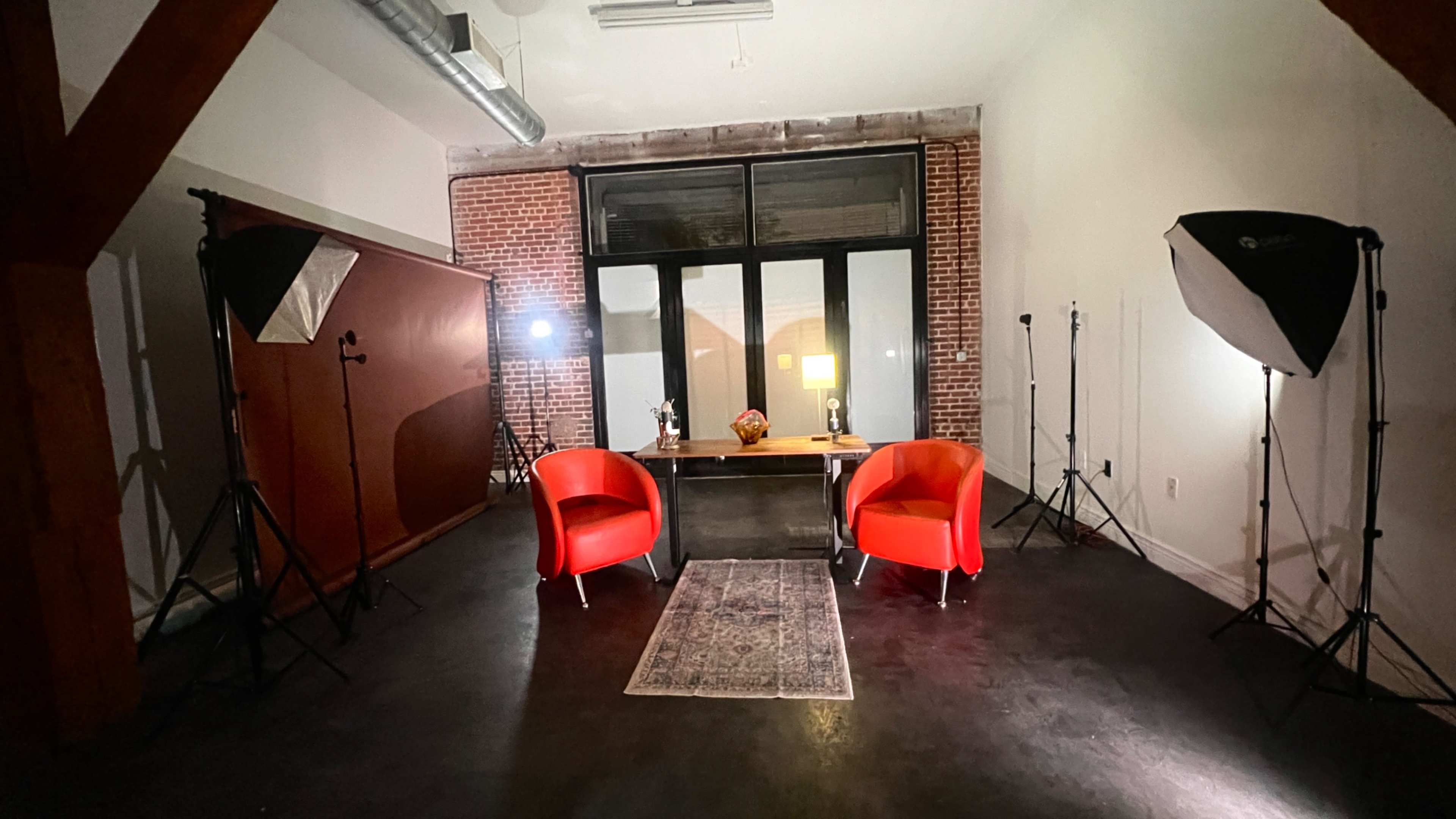 The image shows a minimalist studio setting with two red chairs facing a wooden table, illuminated by softbox lights and featuring a decorative rug on a dark floor.