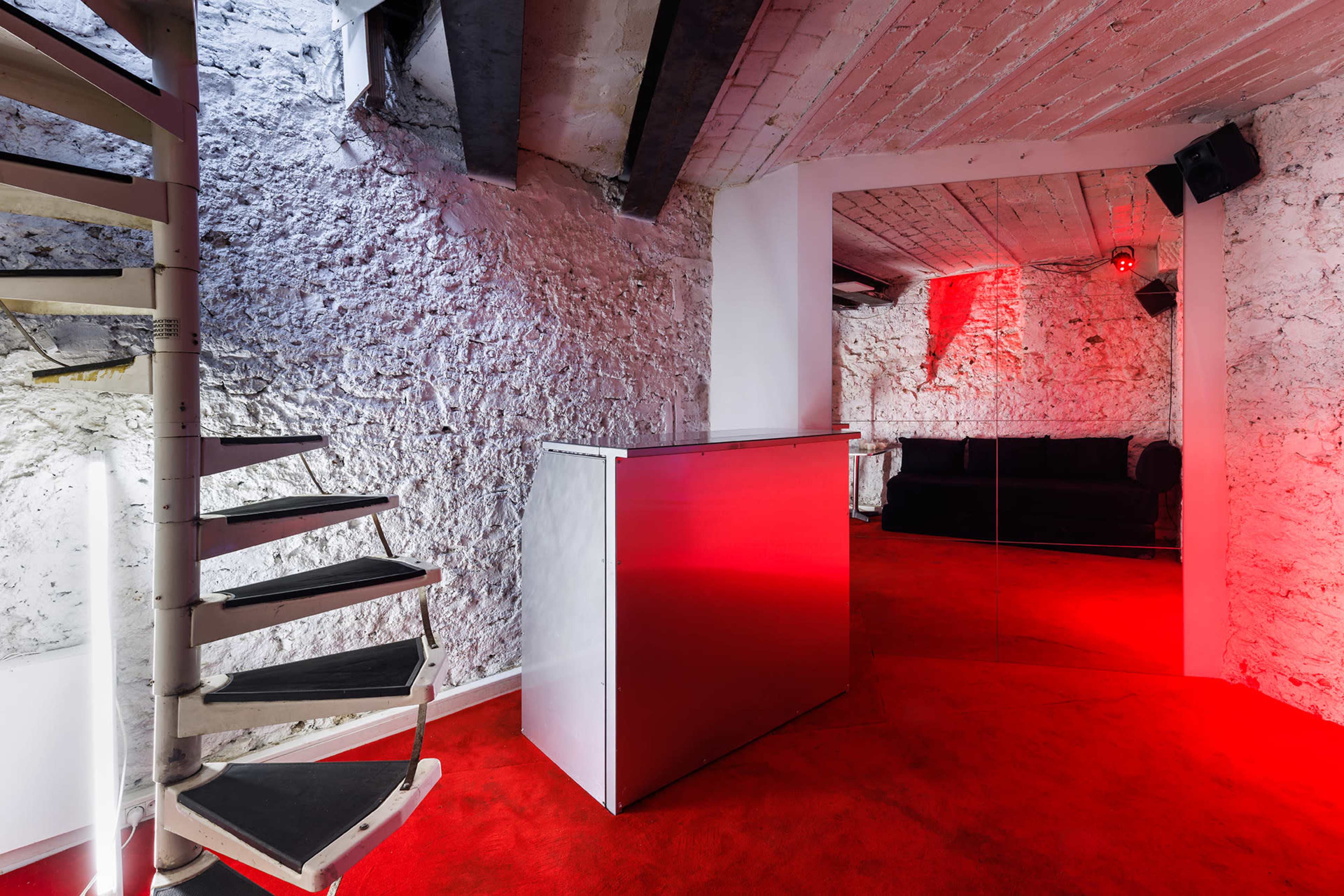 The image shows a modern basement space with white stone walls, a spiral staircase, red carpeting, and red lighting, along with a shiny metallic counter and a black couch.