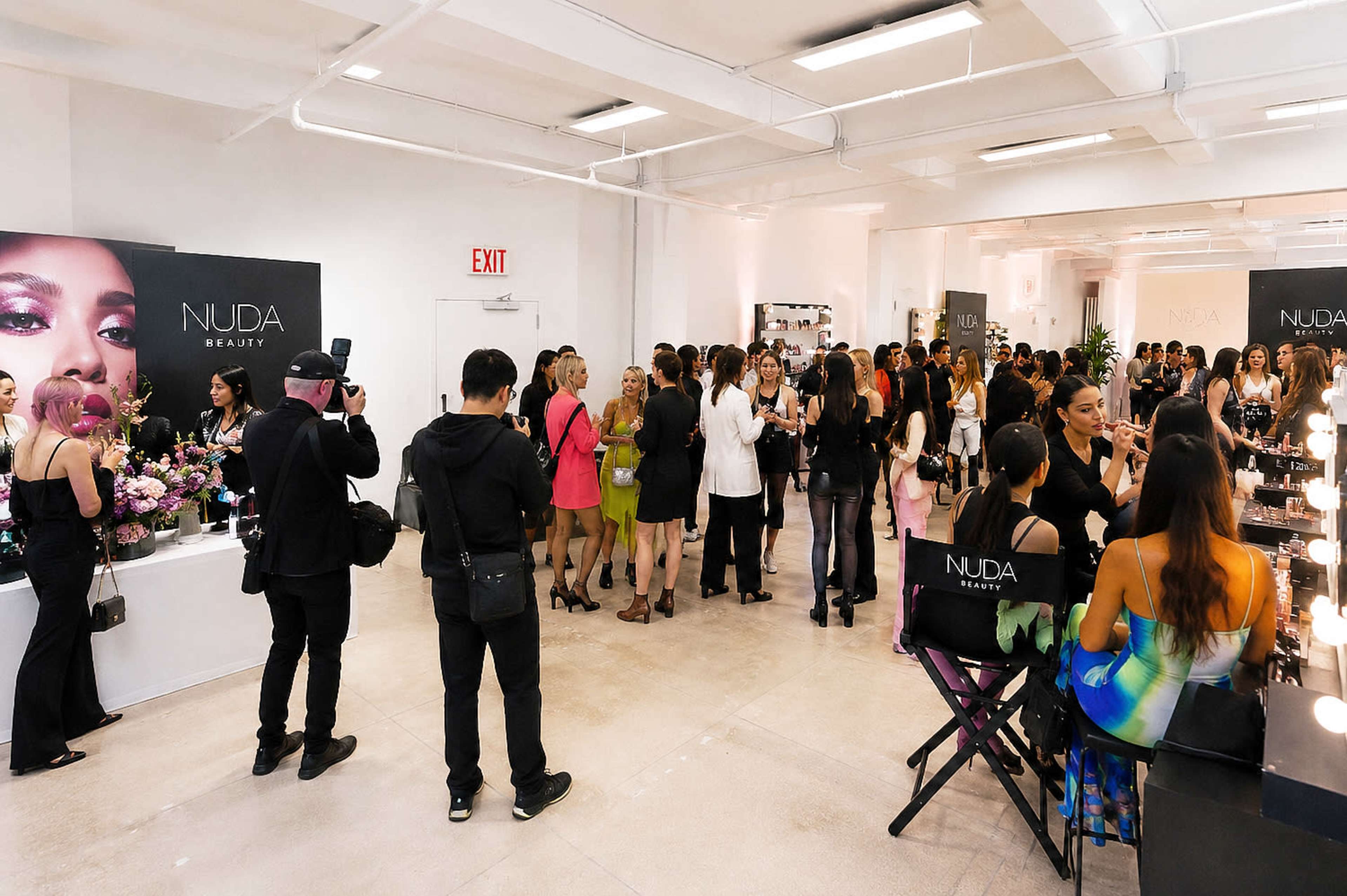 A crowded beauty event features attendees mingling, taking photos, and showcasing products in a well-lit indoor space.