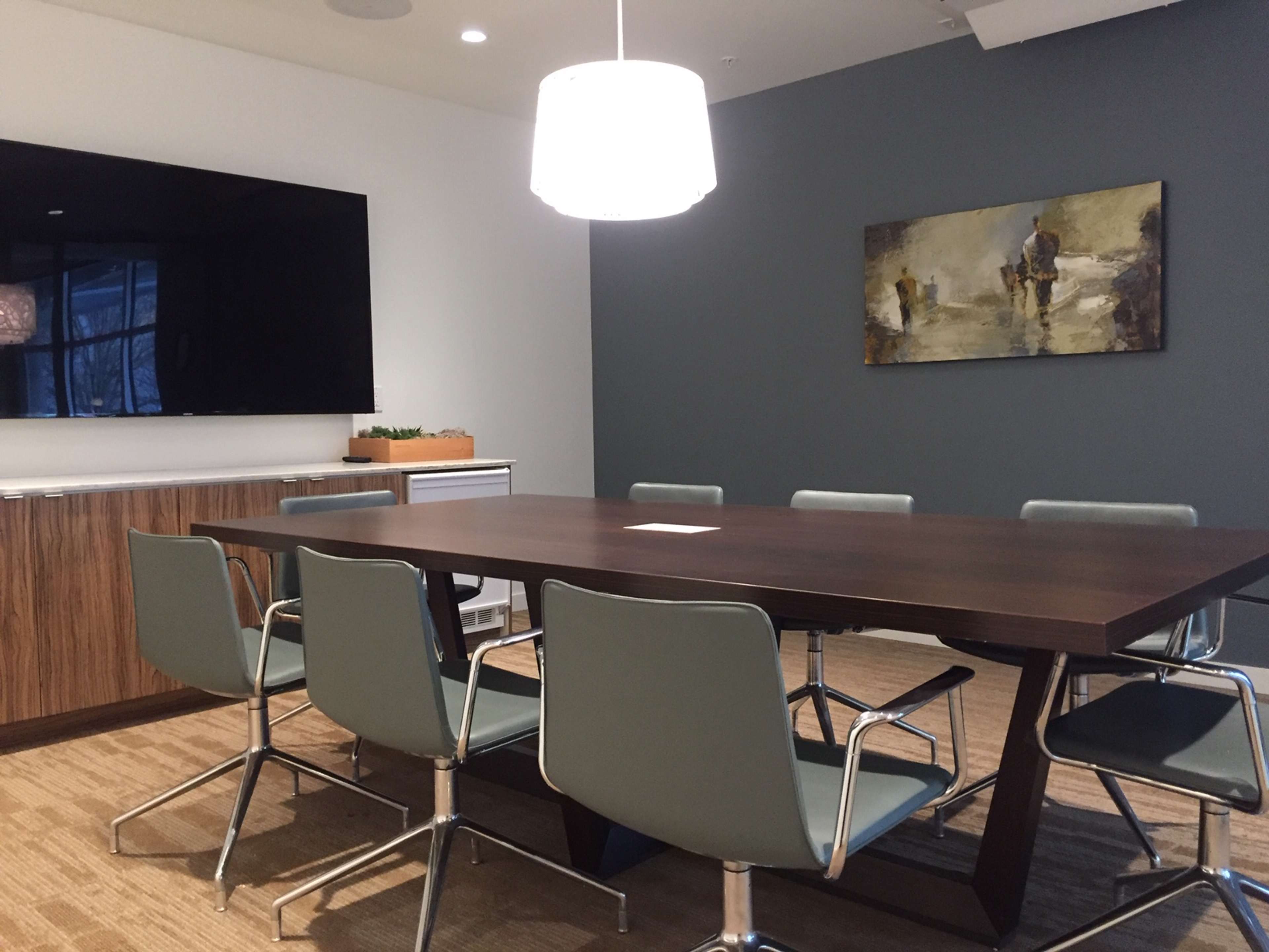 A modern conference room features a large wooden table surrounded by six light blue chairs, a wall-mounted TV, and an abstract painting.