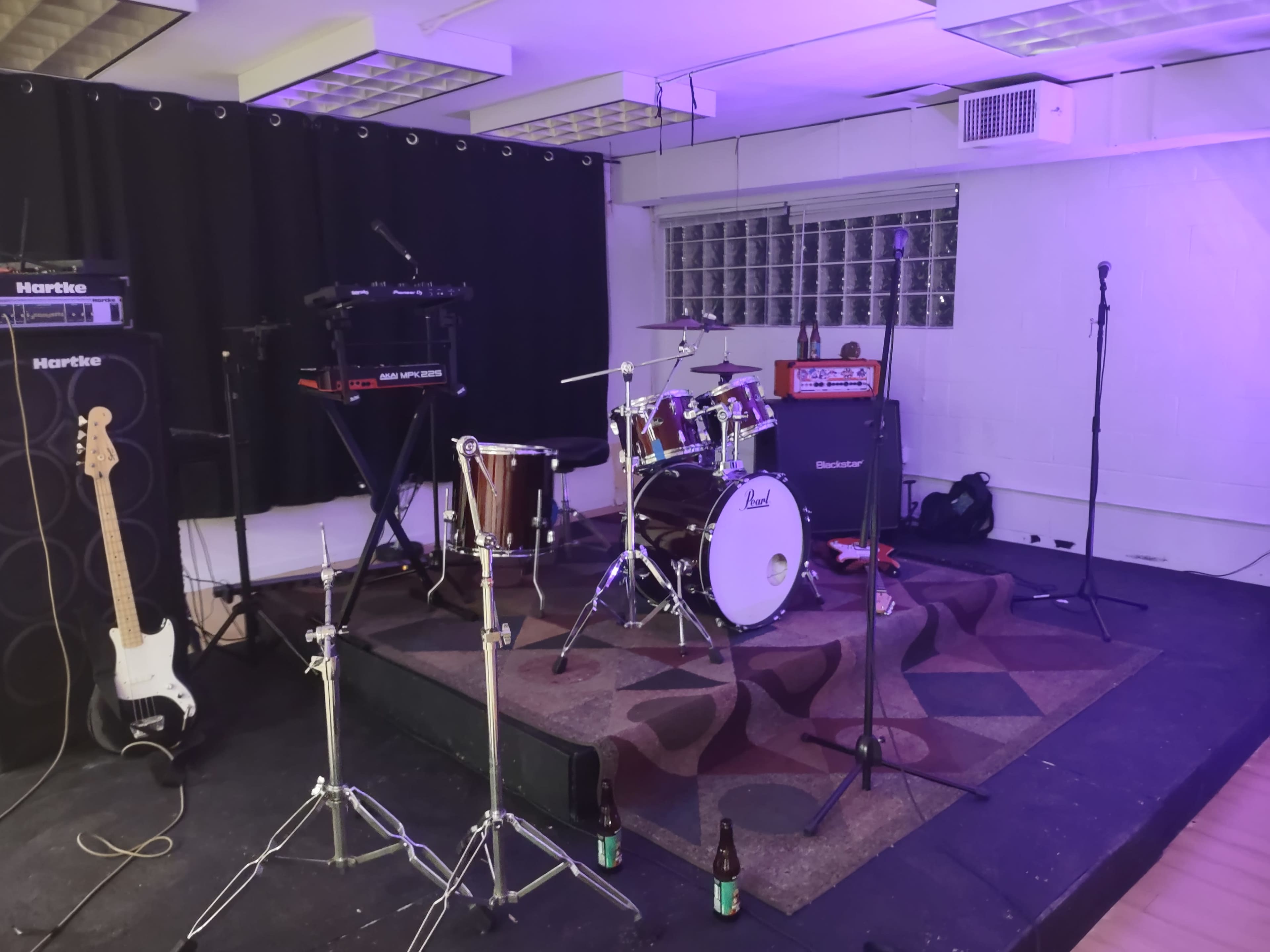 A stage setup with musical instruments, including a drum set, a keyboard, amplifiers, and microphones, in a venue with dim lighting.