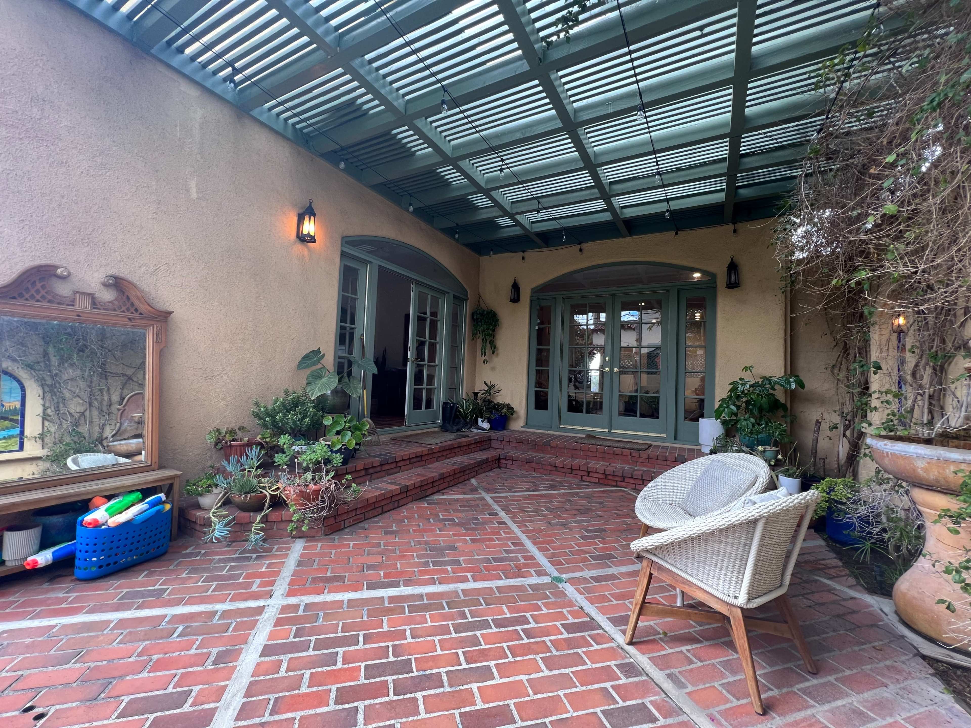 A patio area with brick flooring, potted plants, and a wicker chair, adjacent to a set of glass doors leading into a house.