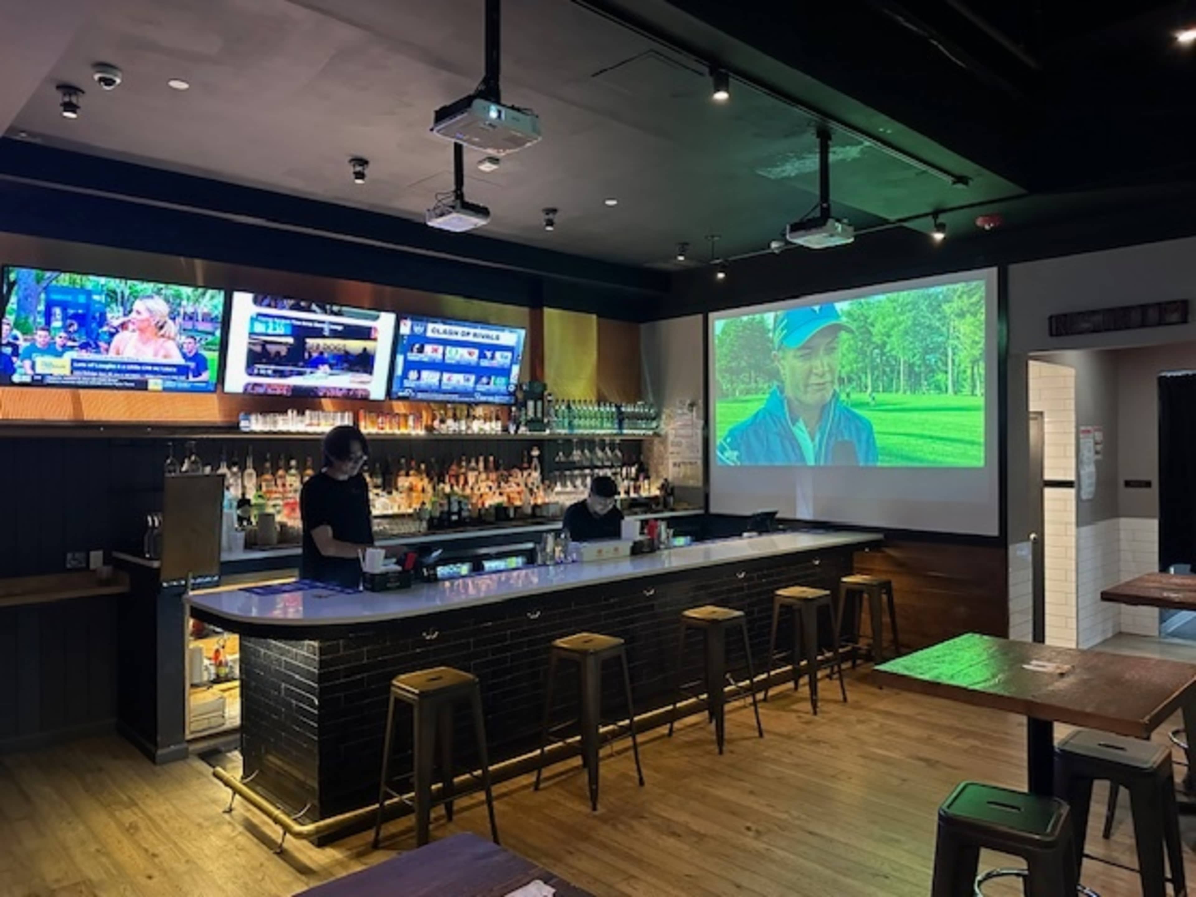 The image shows a modern bar area with multiple flat-screen TVs displaying sports, a large projection screen on one wall, and bar stools arranged around a black counter.