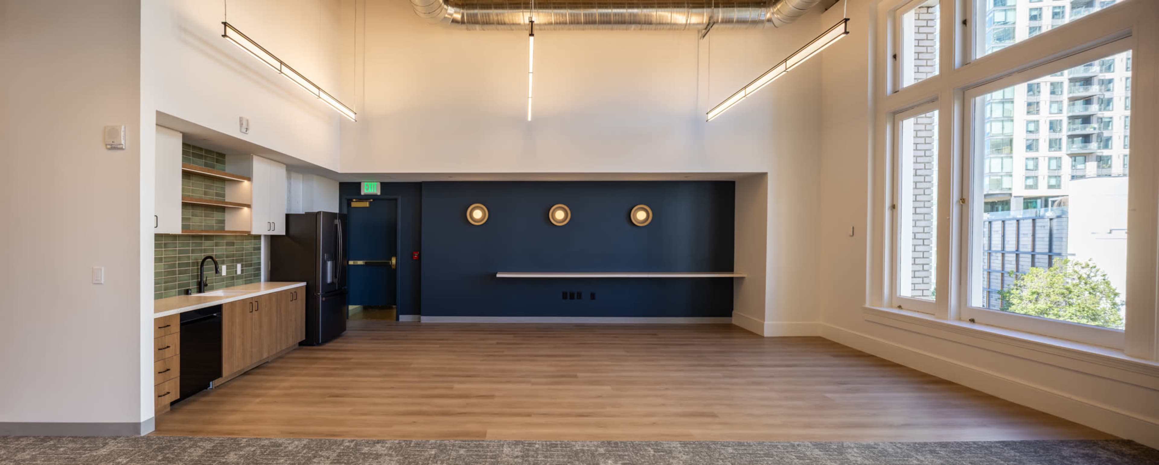 The image shows a spacious, modern room with large windows, a kitchenette area, and a dark accent wall featuring three circular light fixtures.
