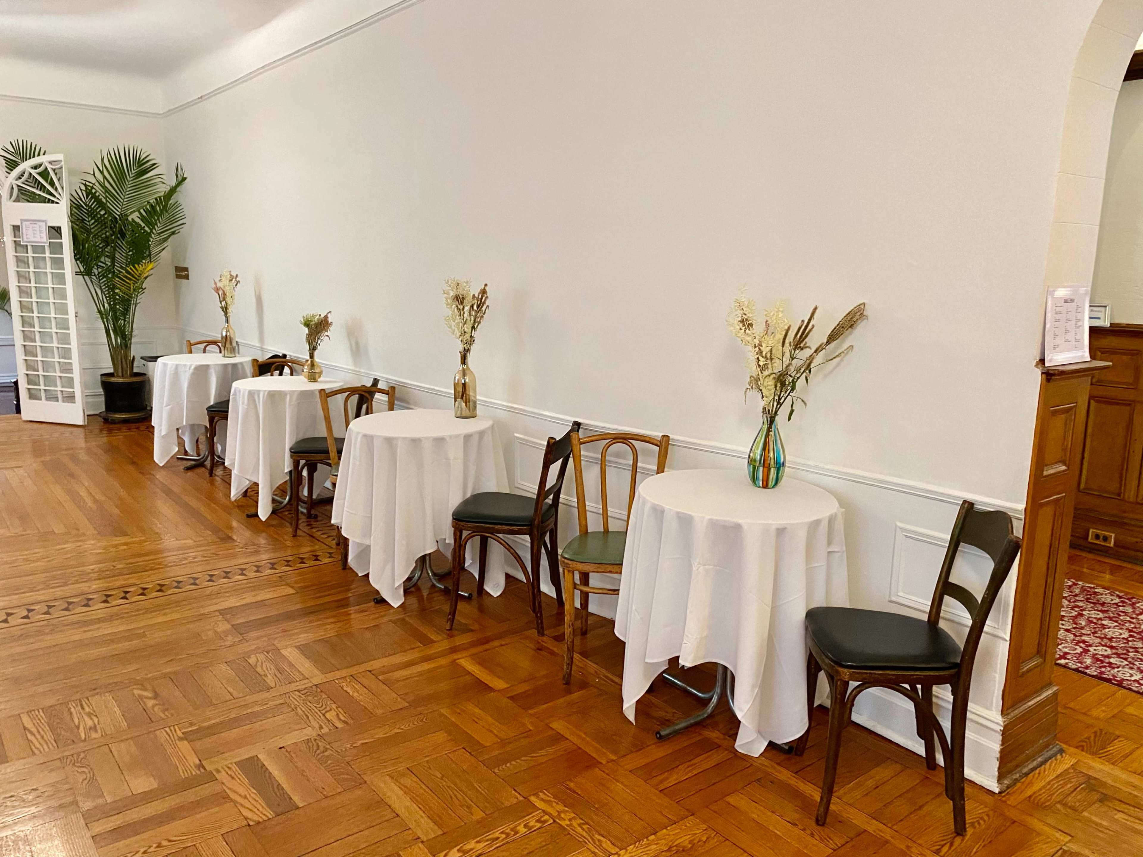 The image shows a row of small tables with white tablecloths and floral centerpieces alongside a wooden floor in a well-lit room.
